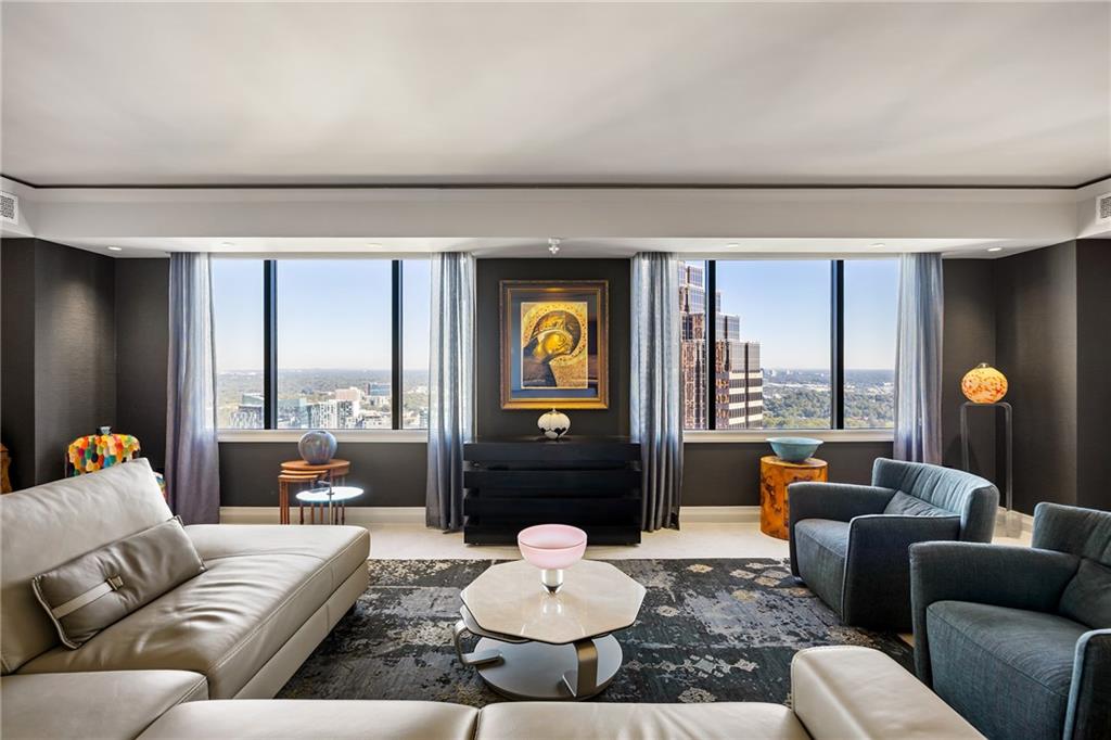 Furnished condo living room with city views through wide windows, leather sofa, blue armchairs, and contemporary art.