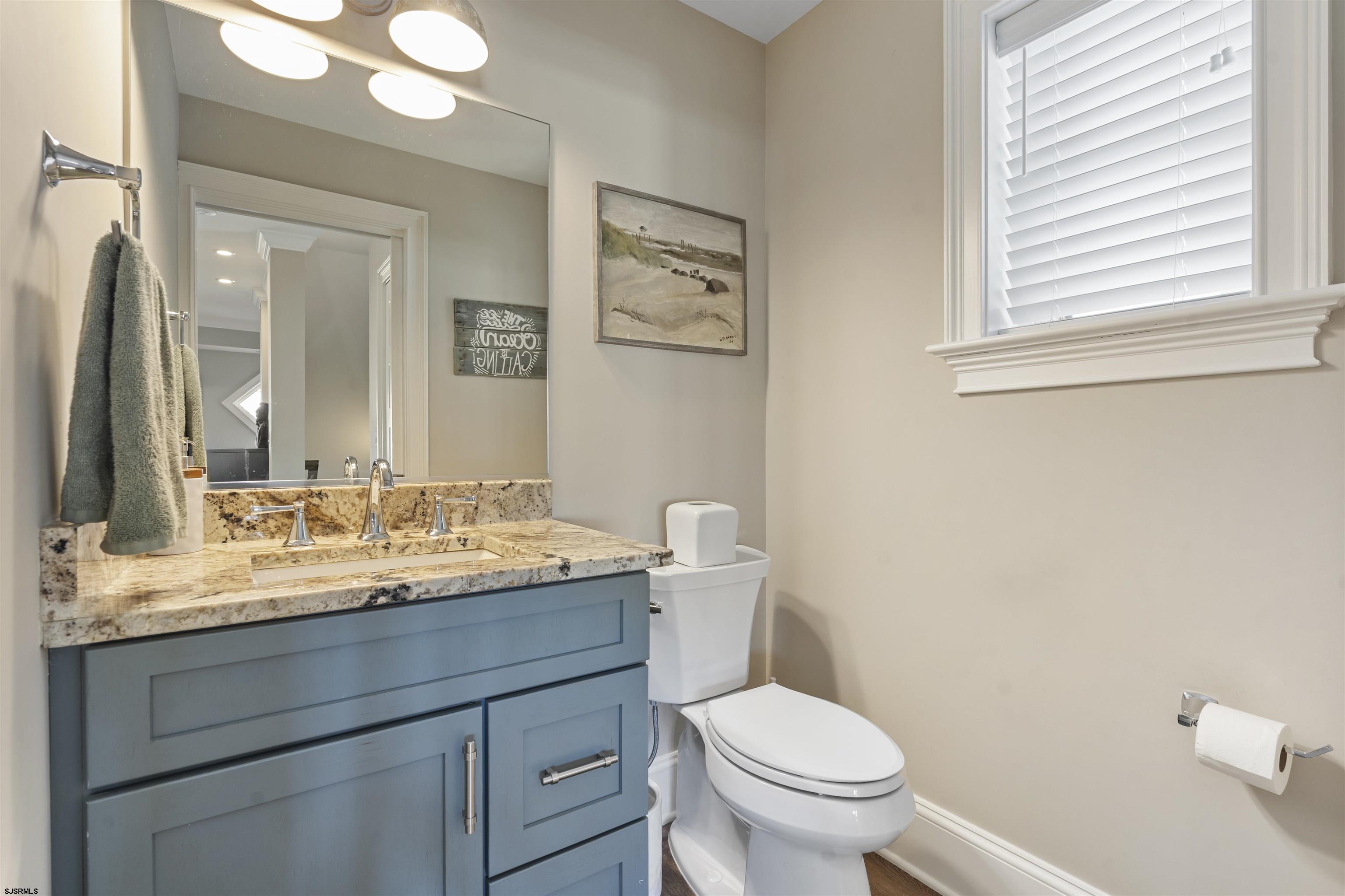 Bathroom with a blue vanity, granite countertop, white toilet, and a window with blinds.