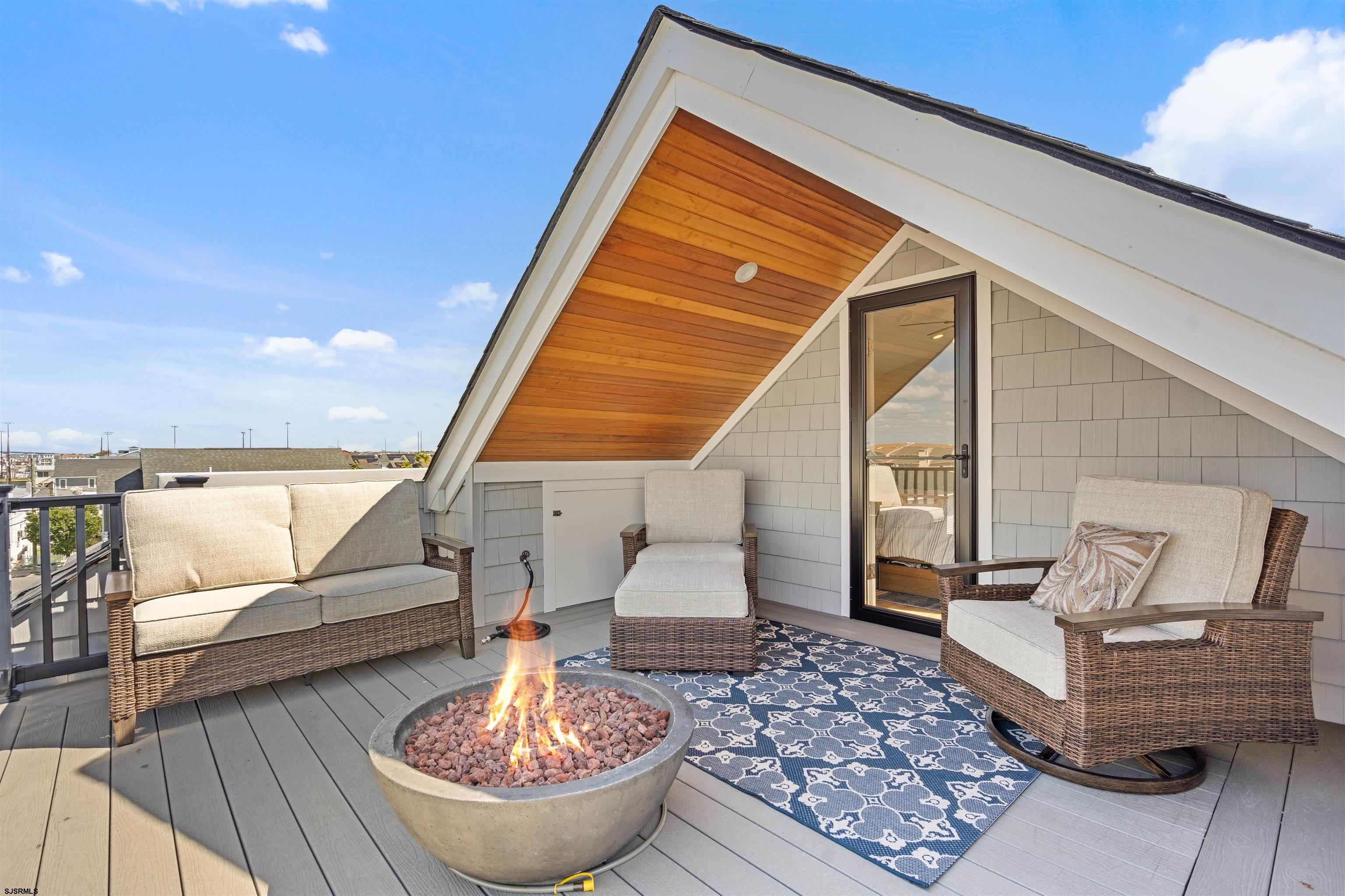 Rooftop deck with wicker seating, a patterned rug, and a lit fire pit beneath a wood-paneled gable.