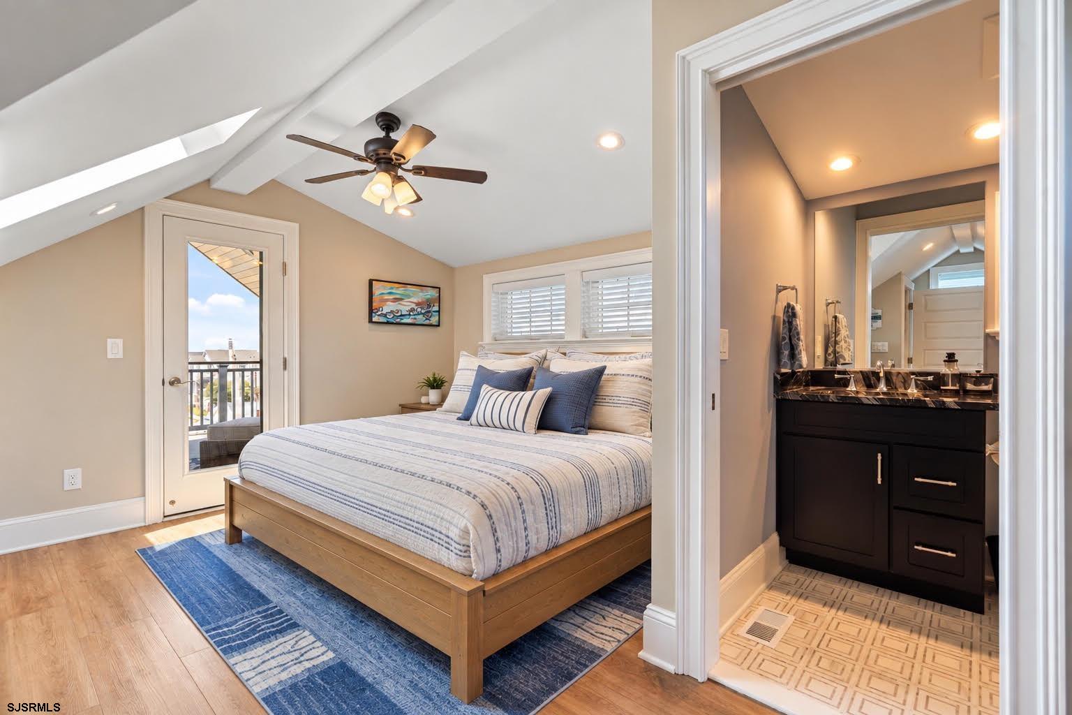 Bedroom with striped bedding and balcony door beside an adjoining bathroom with a dark vanity.
