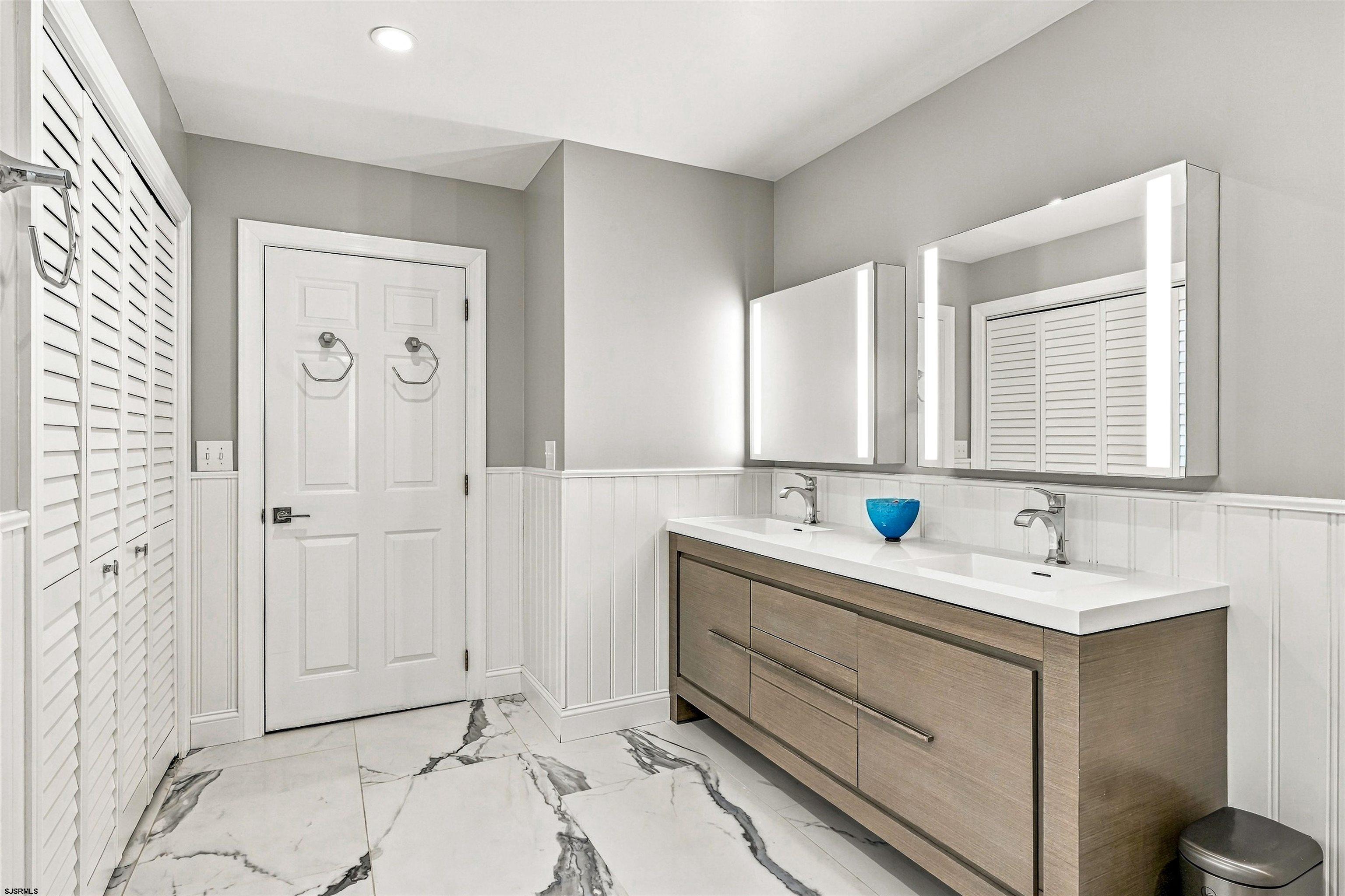 Modern bathroom with a double-sink vanity, illuminated mirrors, and marble-look tile flooring.