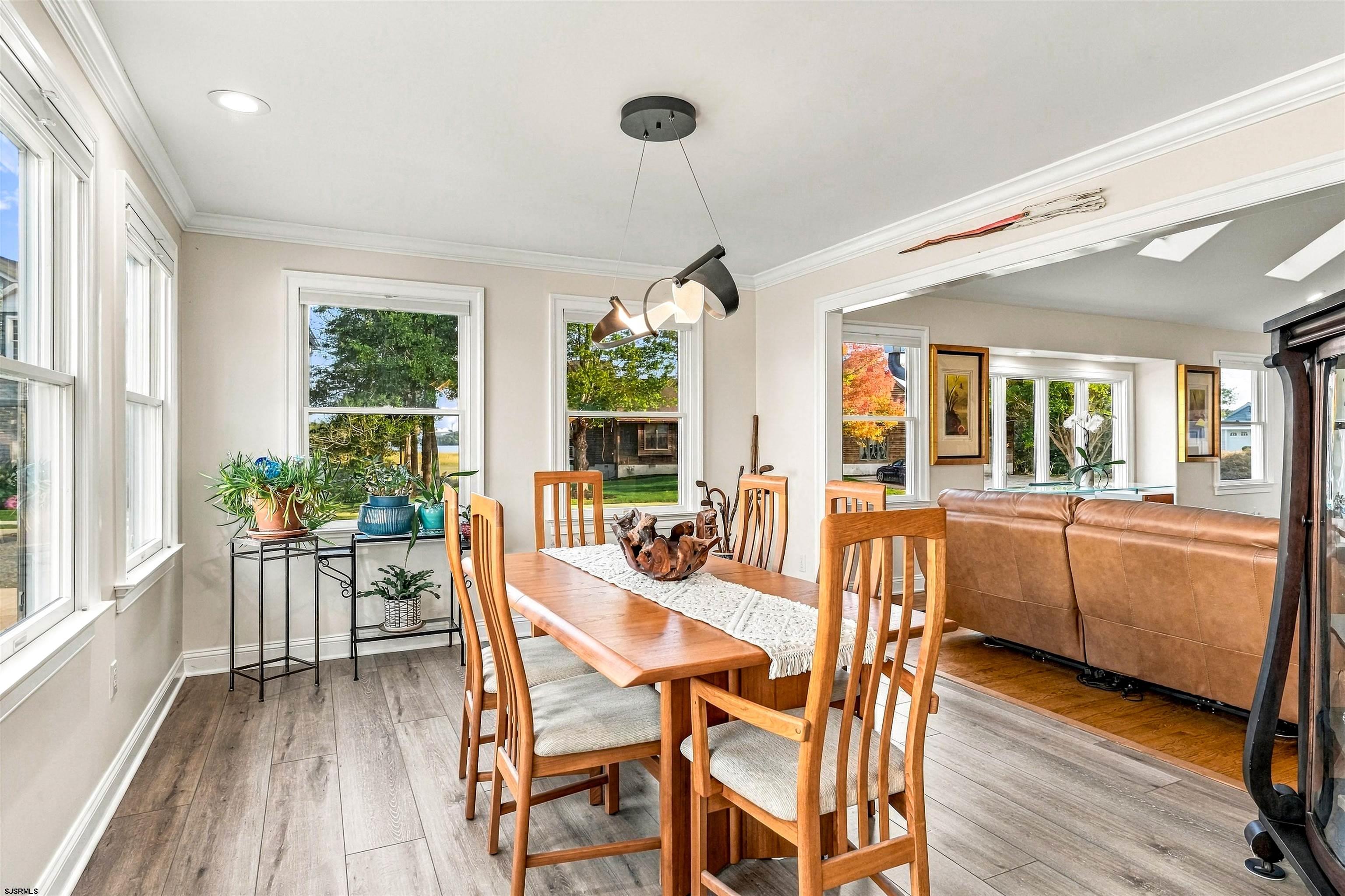 Dining room with a wooden table, large windows, potted plants, and an adjoining living area.