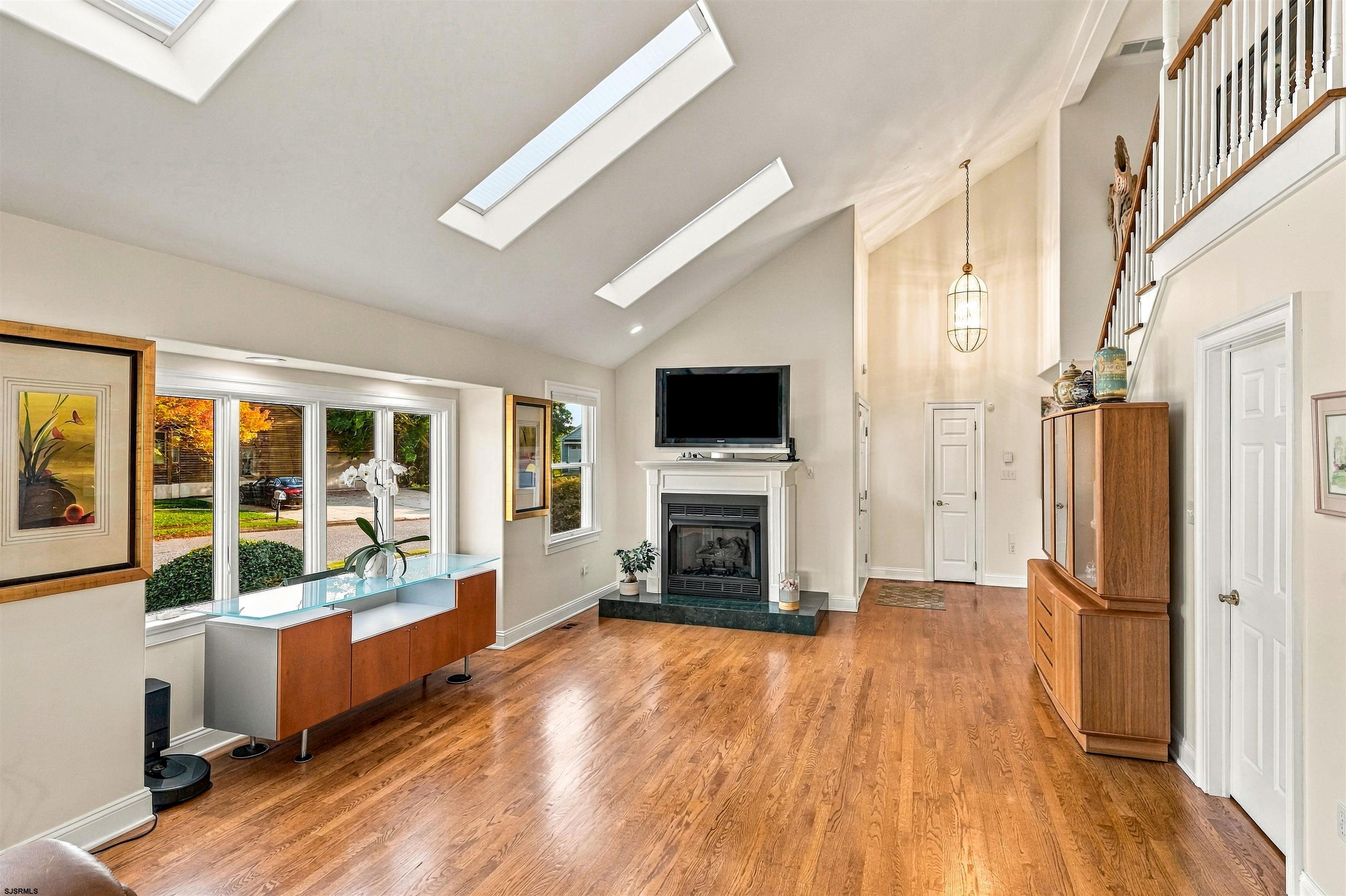 Living room with vaulted skylit ceiling, hardwood floors, fireplace, and large windows.