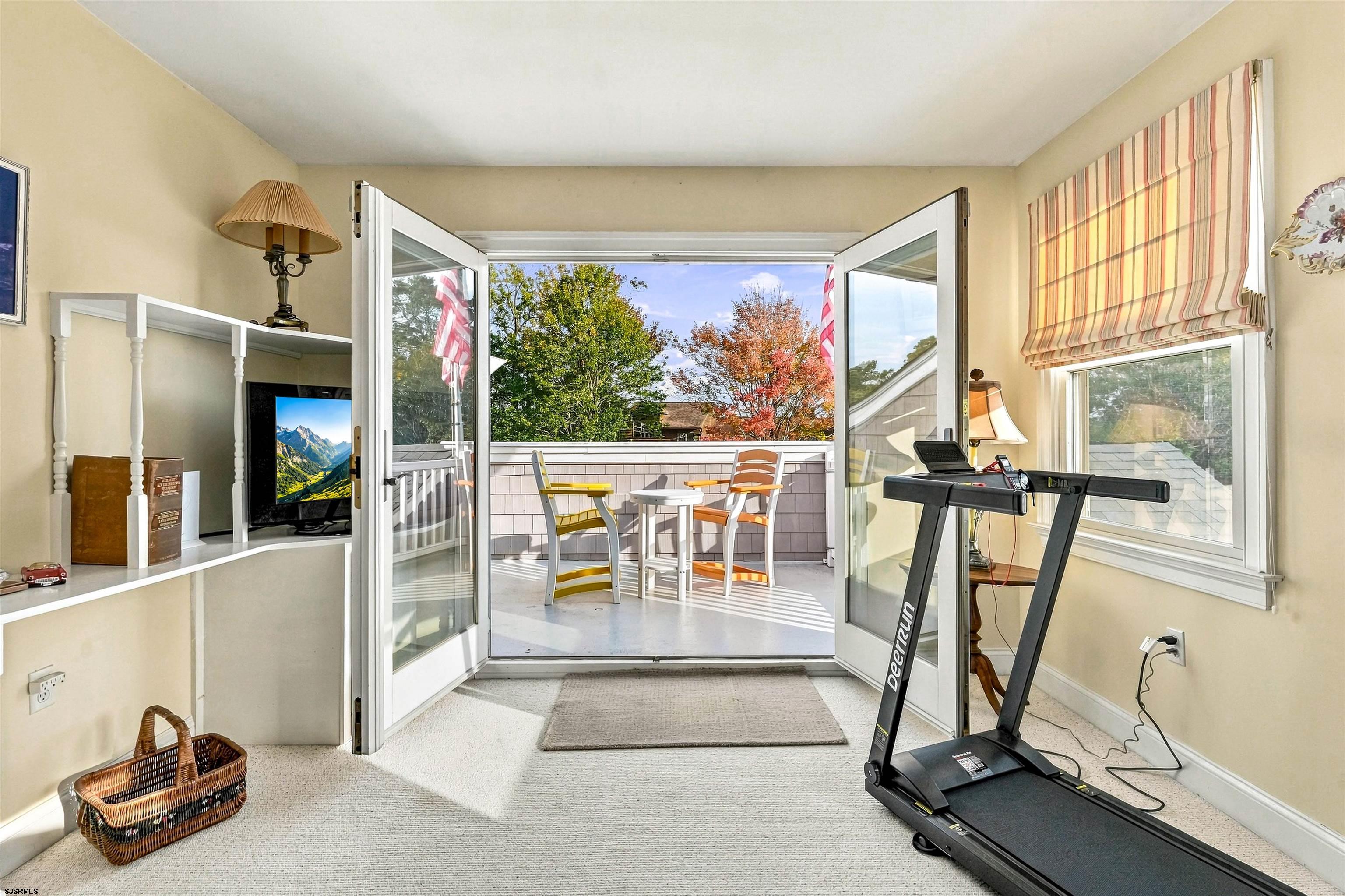 Bright room with open glass doors to a furnished deck, treadmill, and autumn trees beyond.