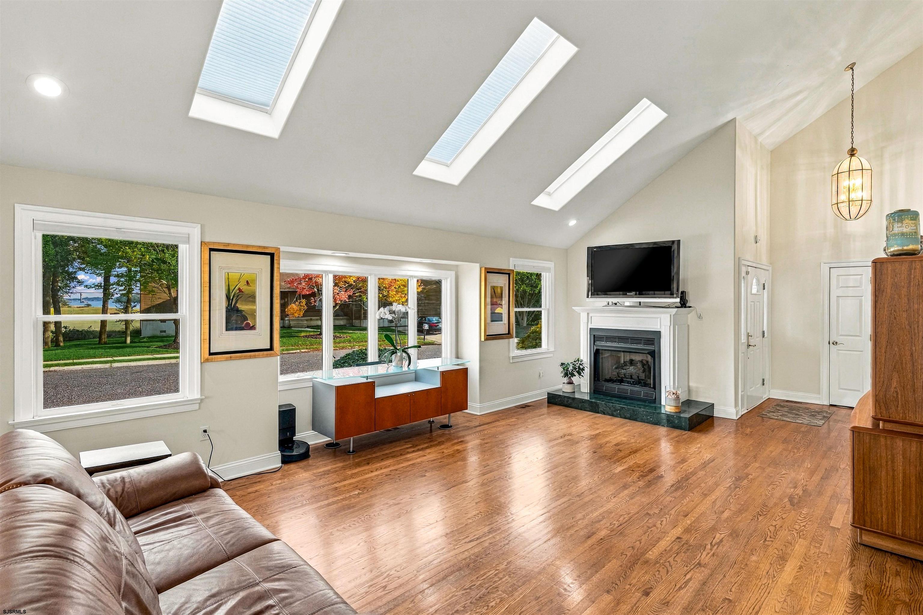 Living room with three skylights, polished hardwood floors, large windows, and a white fireplace.