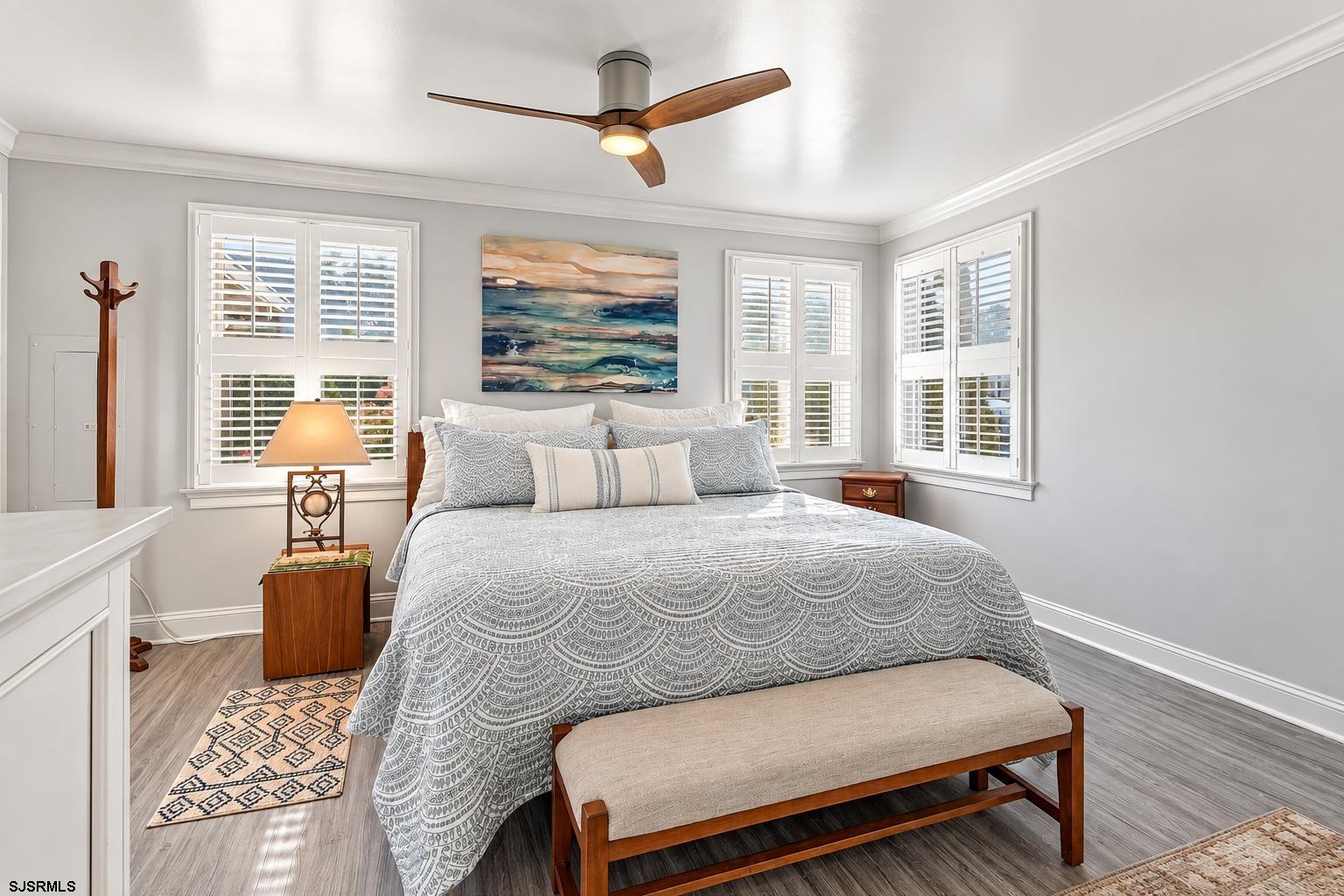 Bedroom with a king-size bed, coastal artwork, and white plantation shutters on three windows.