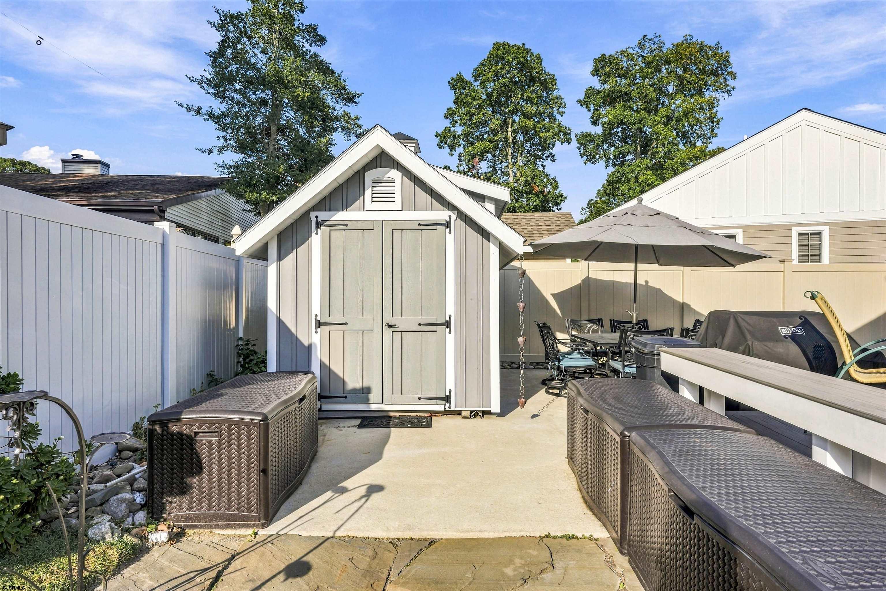 Fenced patio with a white garden shed, wicker benches, and an umbrella-shaded dining set.