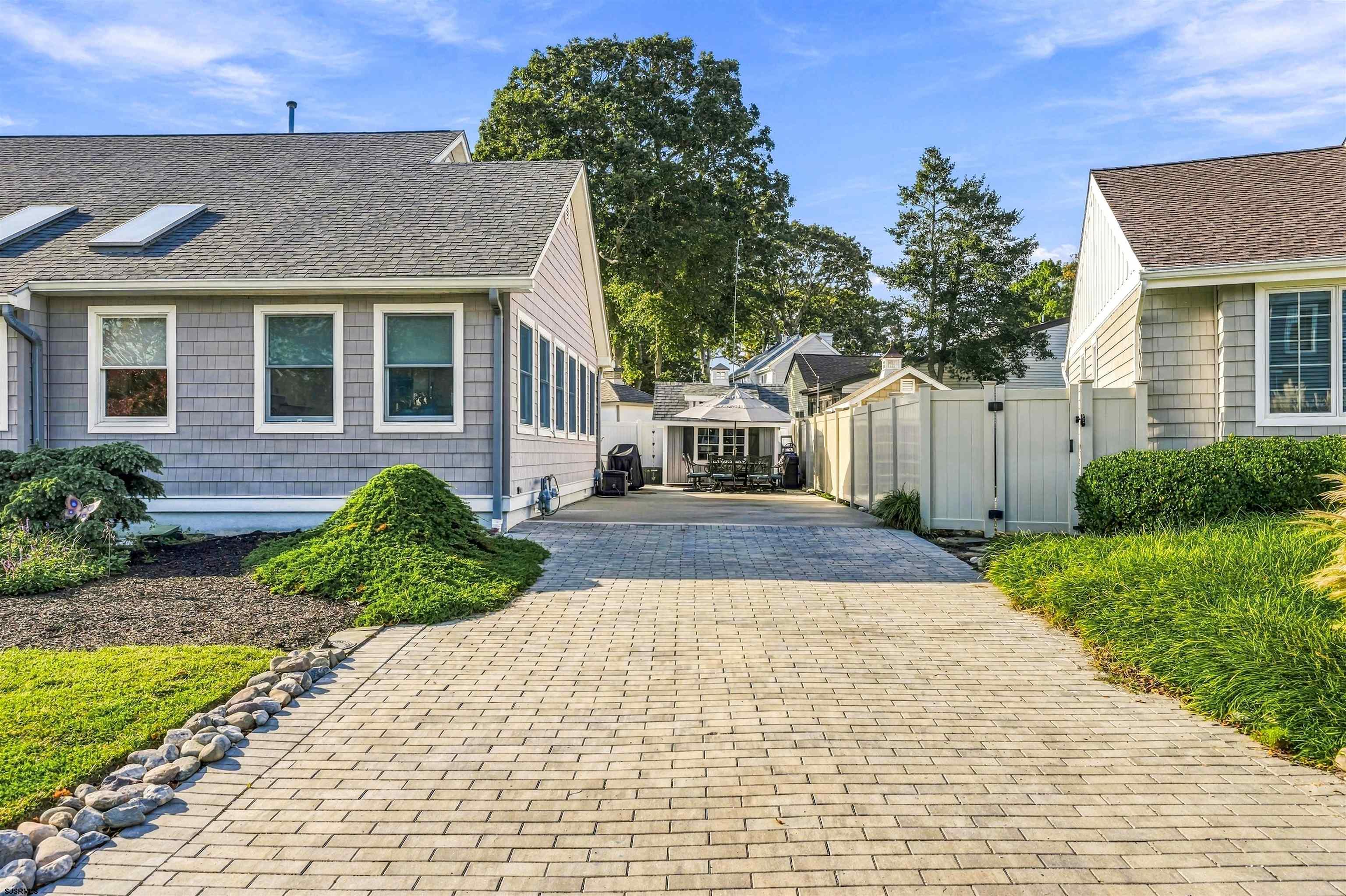 Paver driveway between shingled houses leads to a fenced courtyard with outdoor dining furniture and a pavilion.