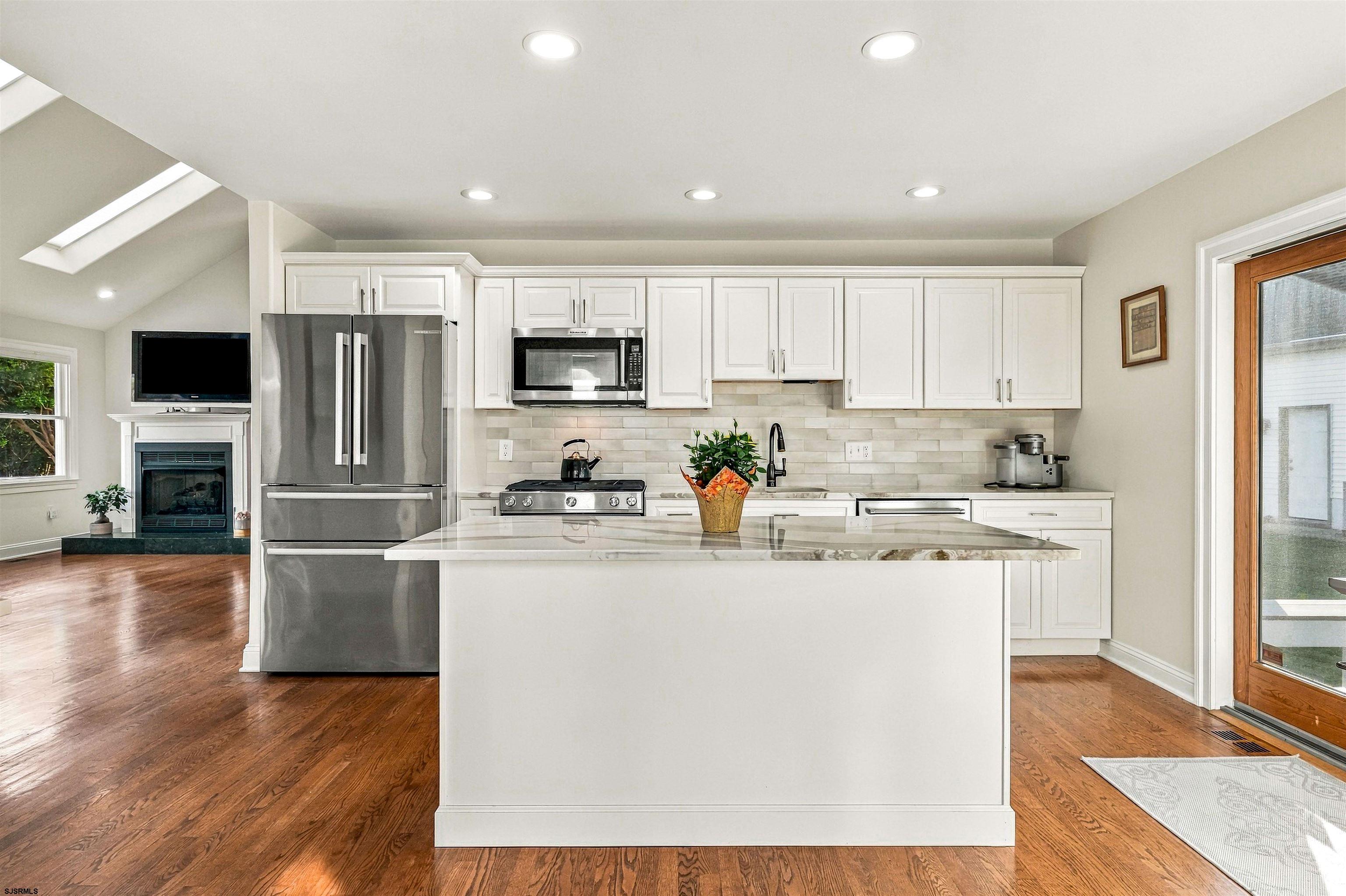 Bright white kitchen with a large stone-topped island, stainless steel appliances, and adjoining fireplace.