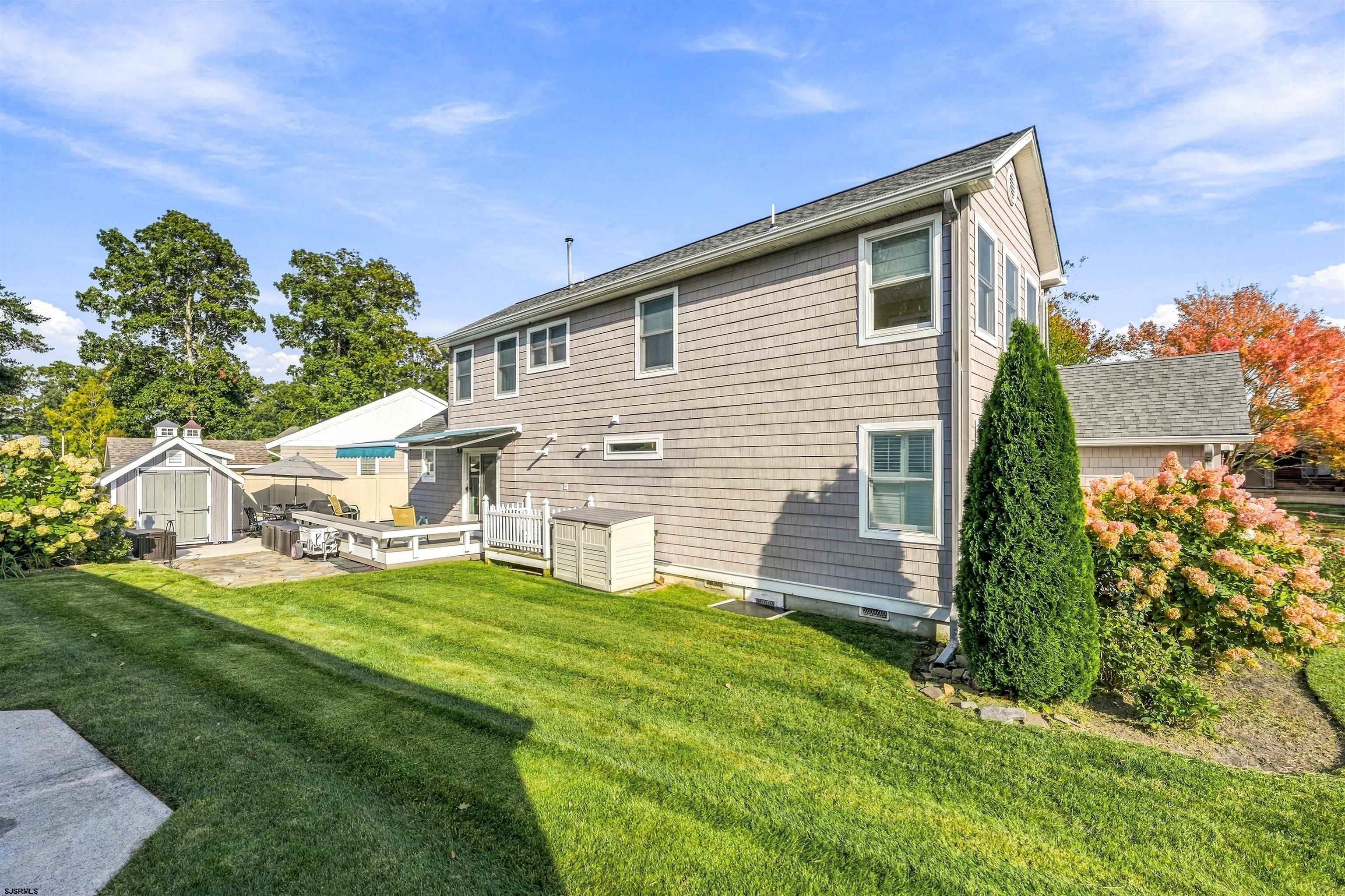 Two-story shingle-sided home with a broad lawn, landscaped garden, patio seating, and a small shed.