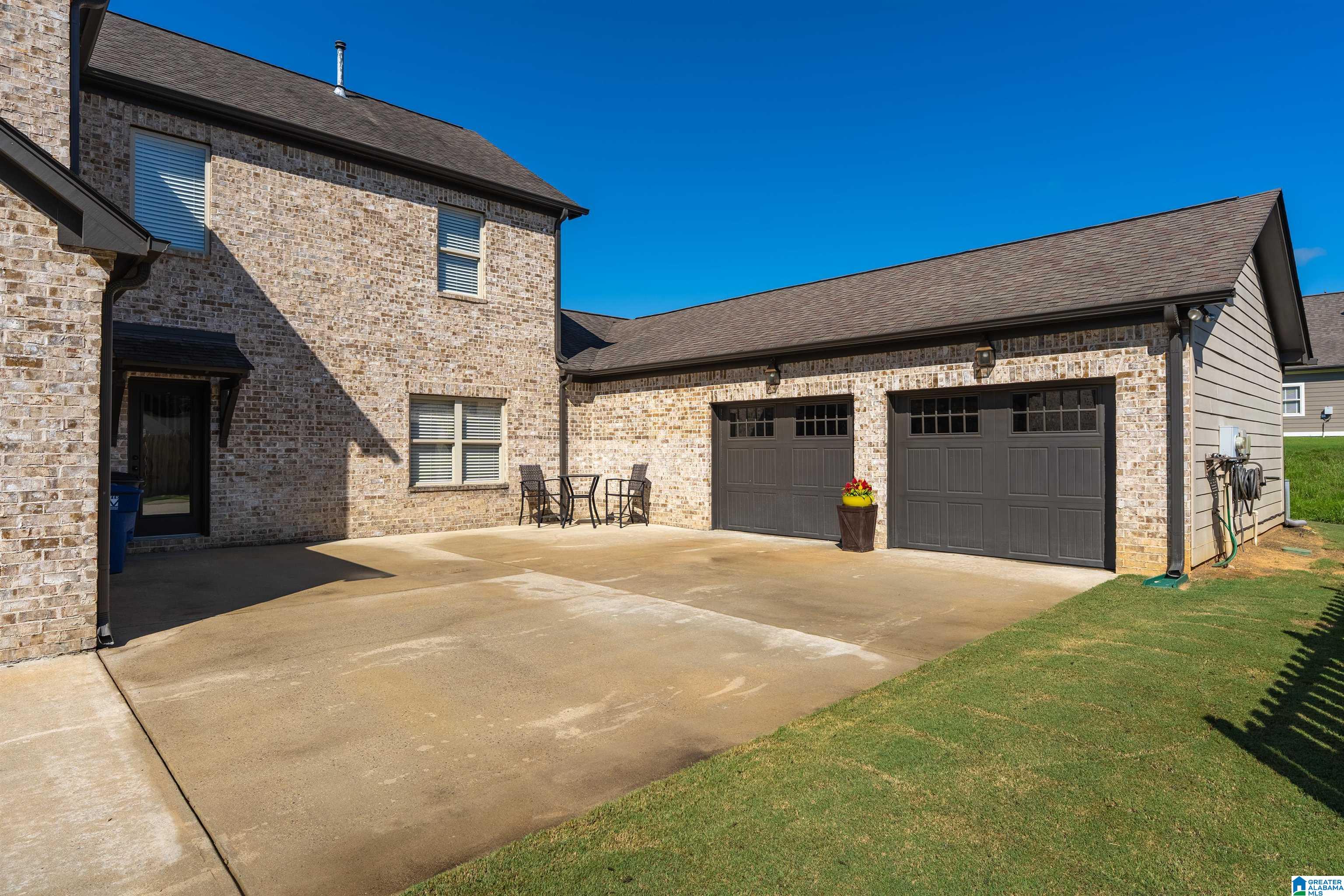 Brick home with two garage doors, a broad concrete driveway, and a small patio seating area.