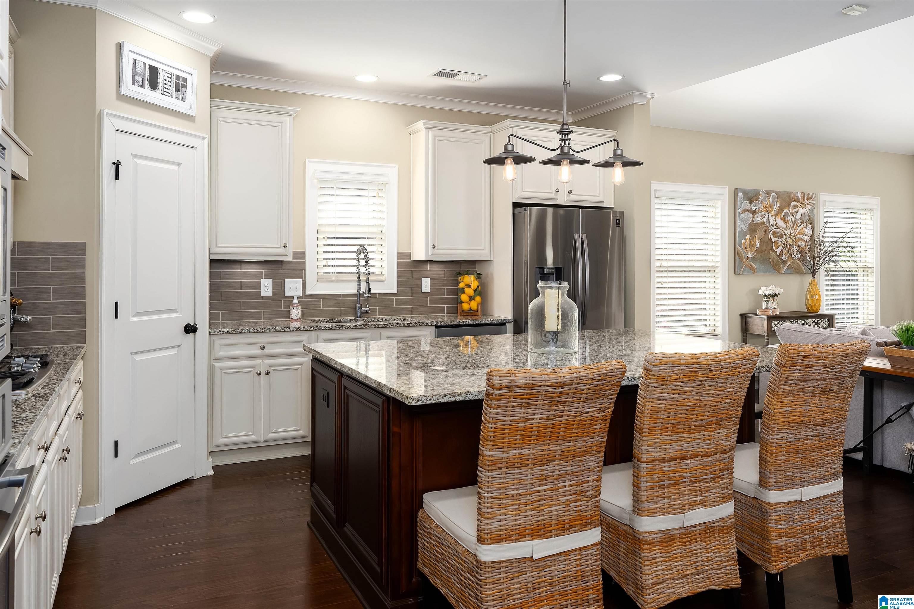 Open-plan kitchen with a granite island, woven bar stools, white cabinetry, and stainless steel refrigerator.