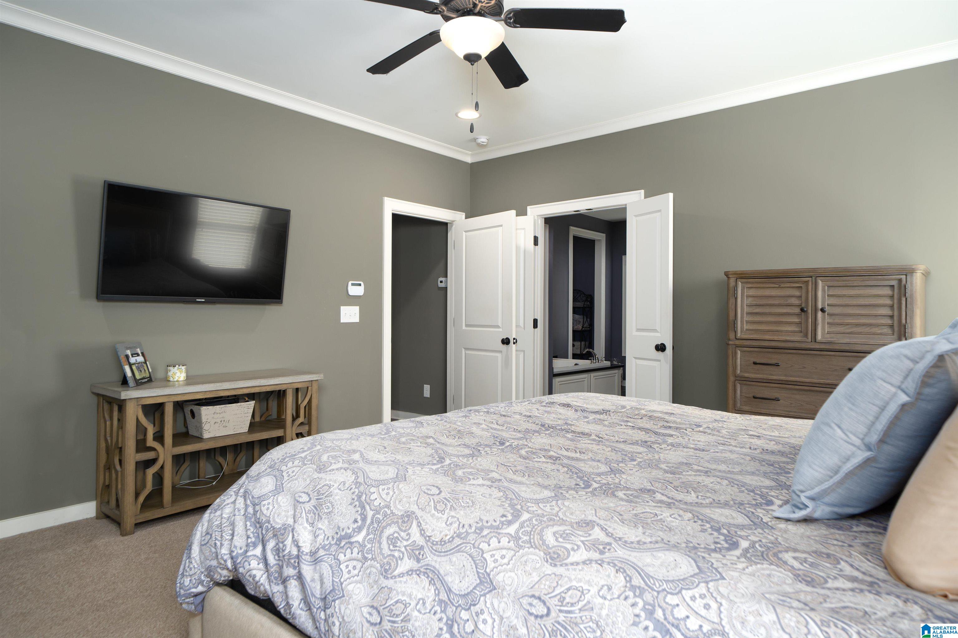 Furnished bedroom with a patterned bedspread, ceiling fan, wall-mounted television, and open bathroom doors.