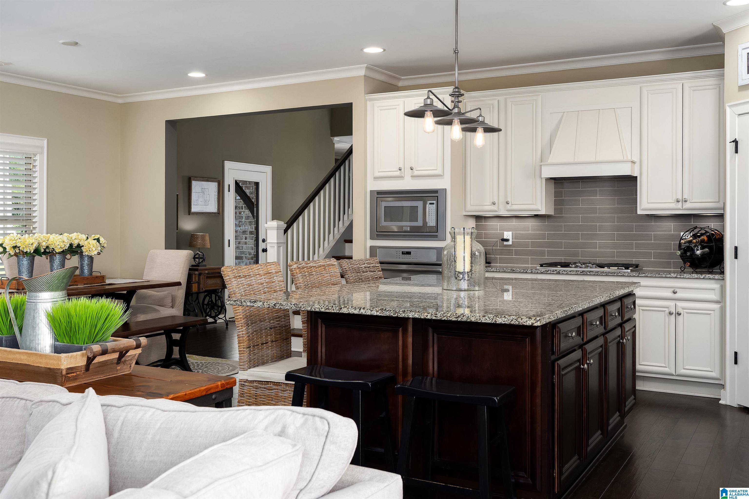 Open-plan kitchen with a granite island, dark wood cabinetry, white cabinets, and gray tile backsplash.