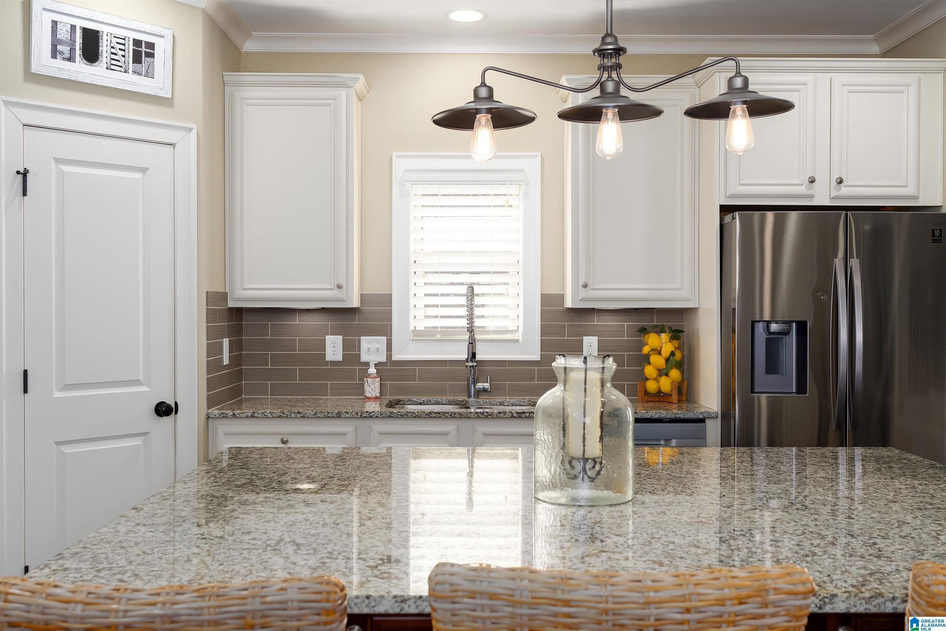 Kitchen with a granite island, wicker stools, white cabinets, and stainless steel refrigerator.