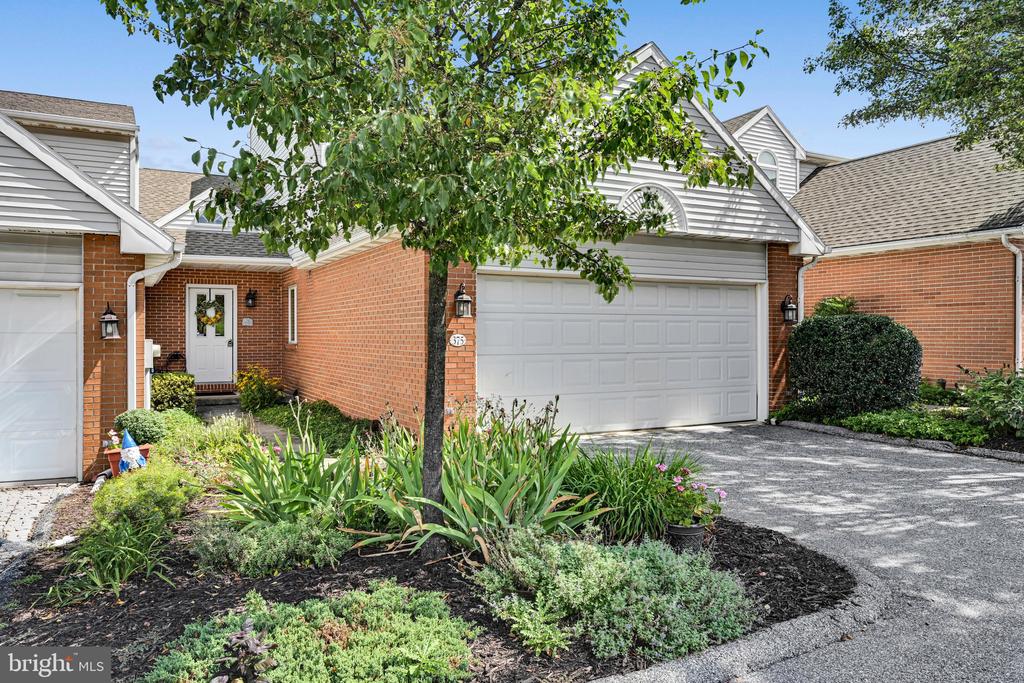 Brick townhouse with a white upper facade, two-car garage, and landscaped garden shaded by a leafy tree.