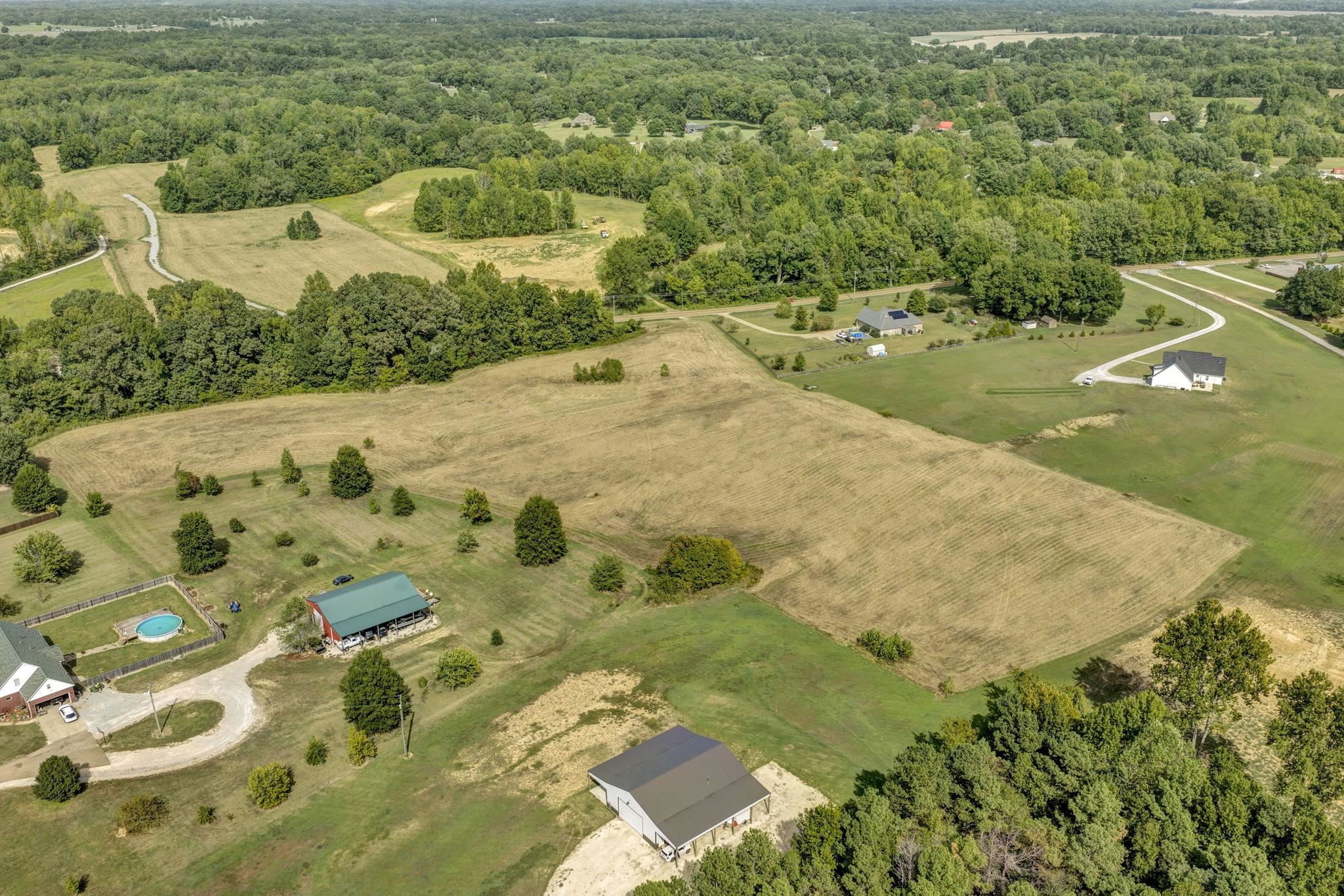 Aerial view of open acreage bordered by woods, with nearby homes and farm buildings.