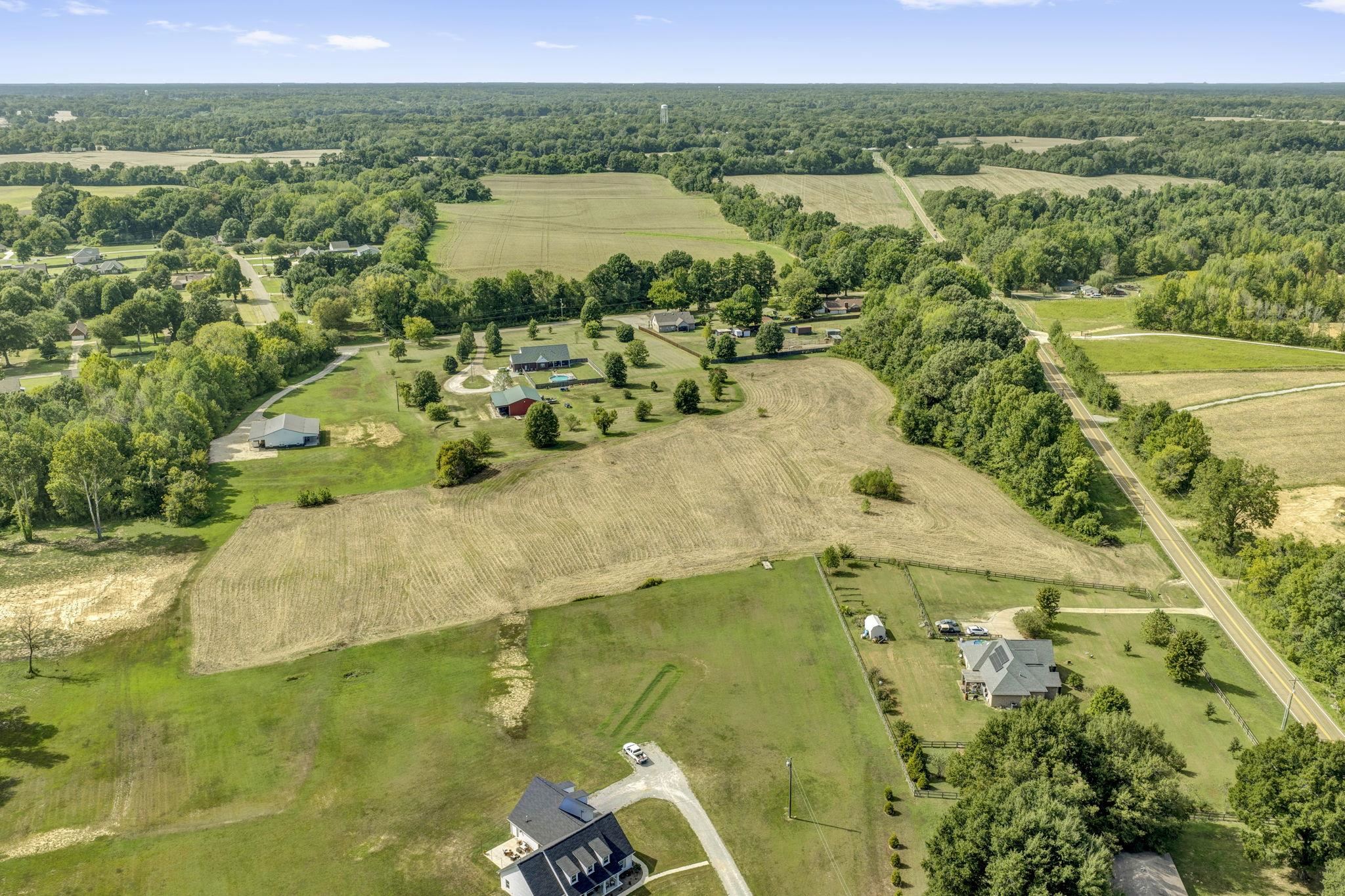 Aerial view of rural fields bordered by trees, neighboring homes, and a paved road.