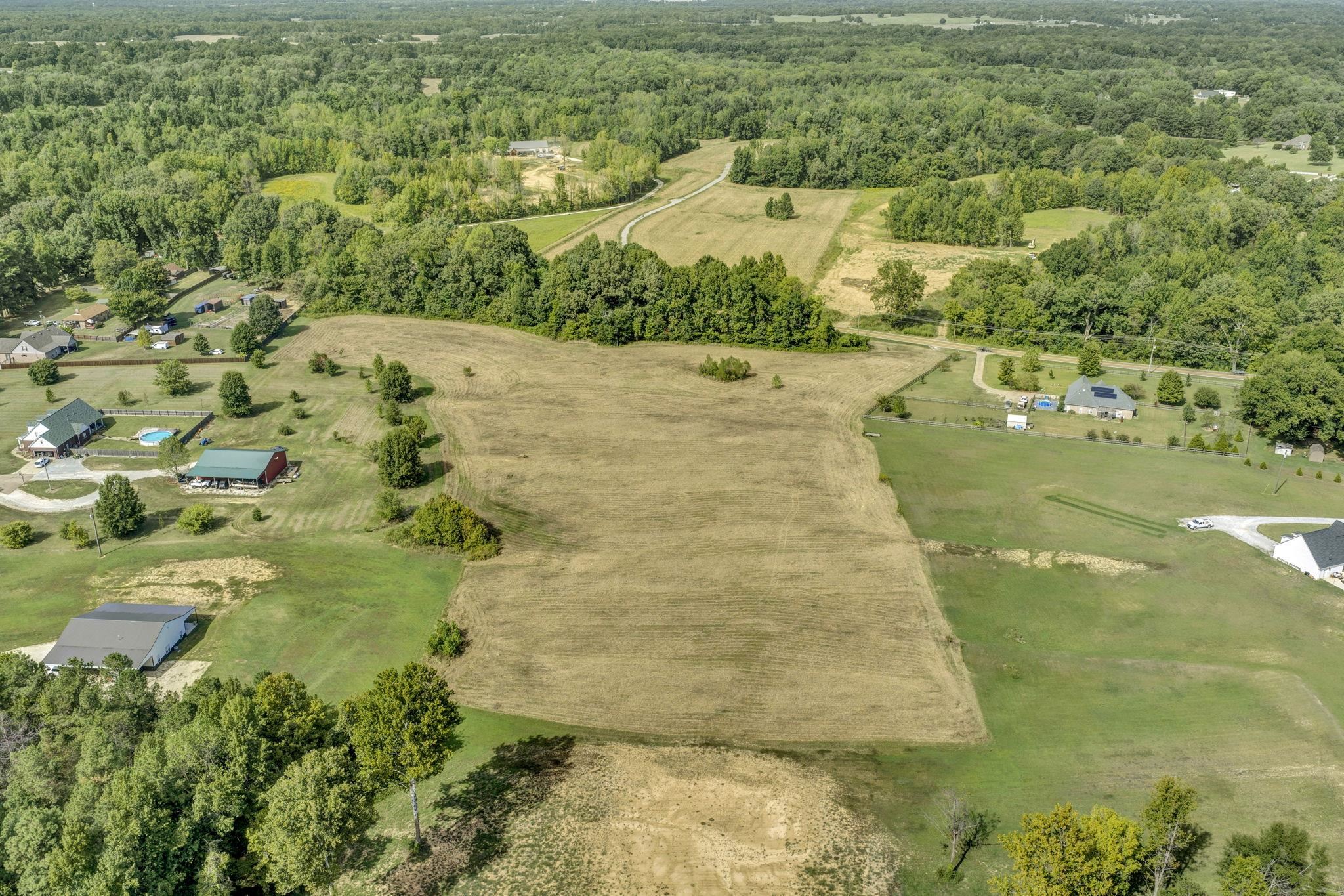 Aerial view of a broad cleared parcel bordered by mature trees, with neighboring homes and a road nearby.