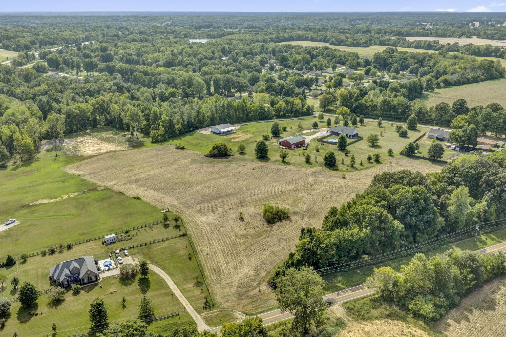 Aerial view of open rural land with a broad harvested field and wooded borders beside a road.