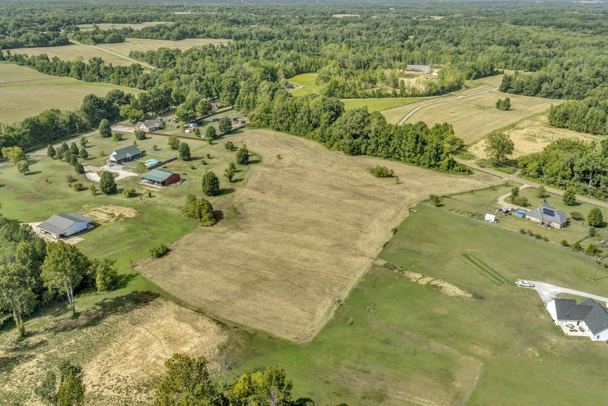 Aerial view of a broad vacant field bordered by trees, lawns, and neighboring homes.