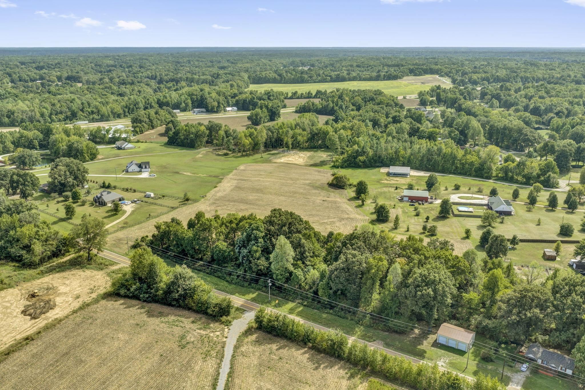 Aerial view of open rural lots bordered by trees, with nearby homes and a road.