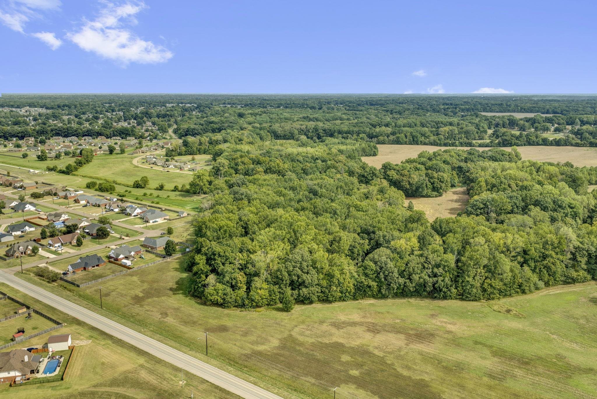 Aerial view of wooded acreage and open fields beside a road and neighborhood.