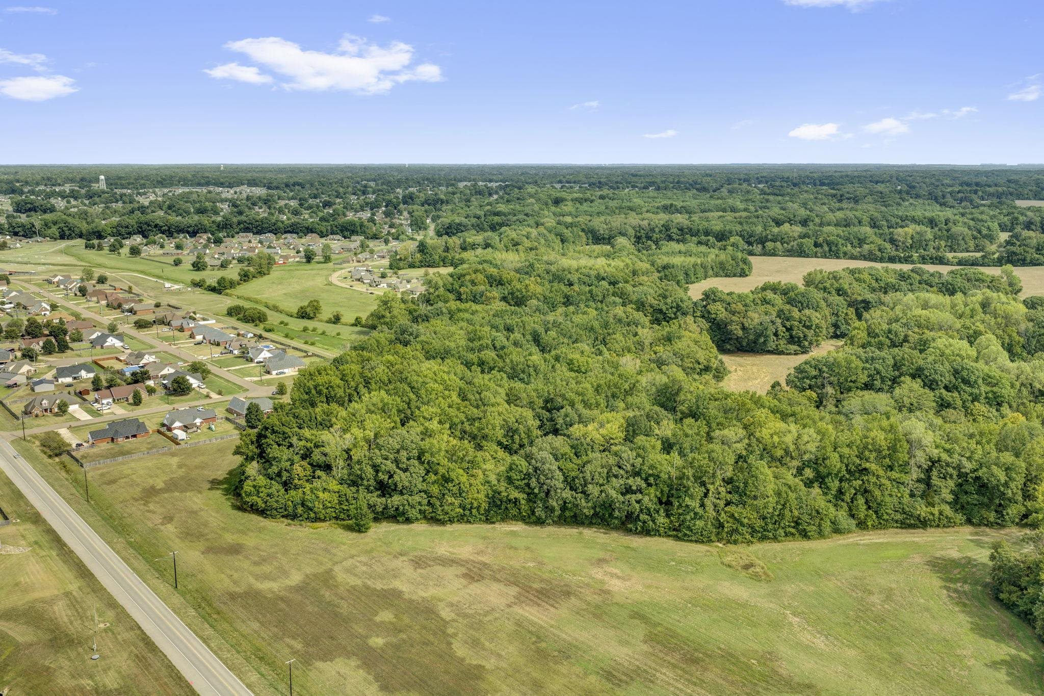 Aerial view of wooded land and open fields beside a road, with a neighborhood nearby.