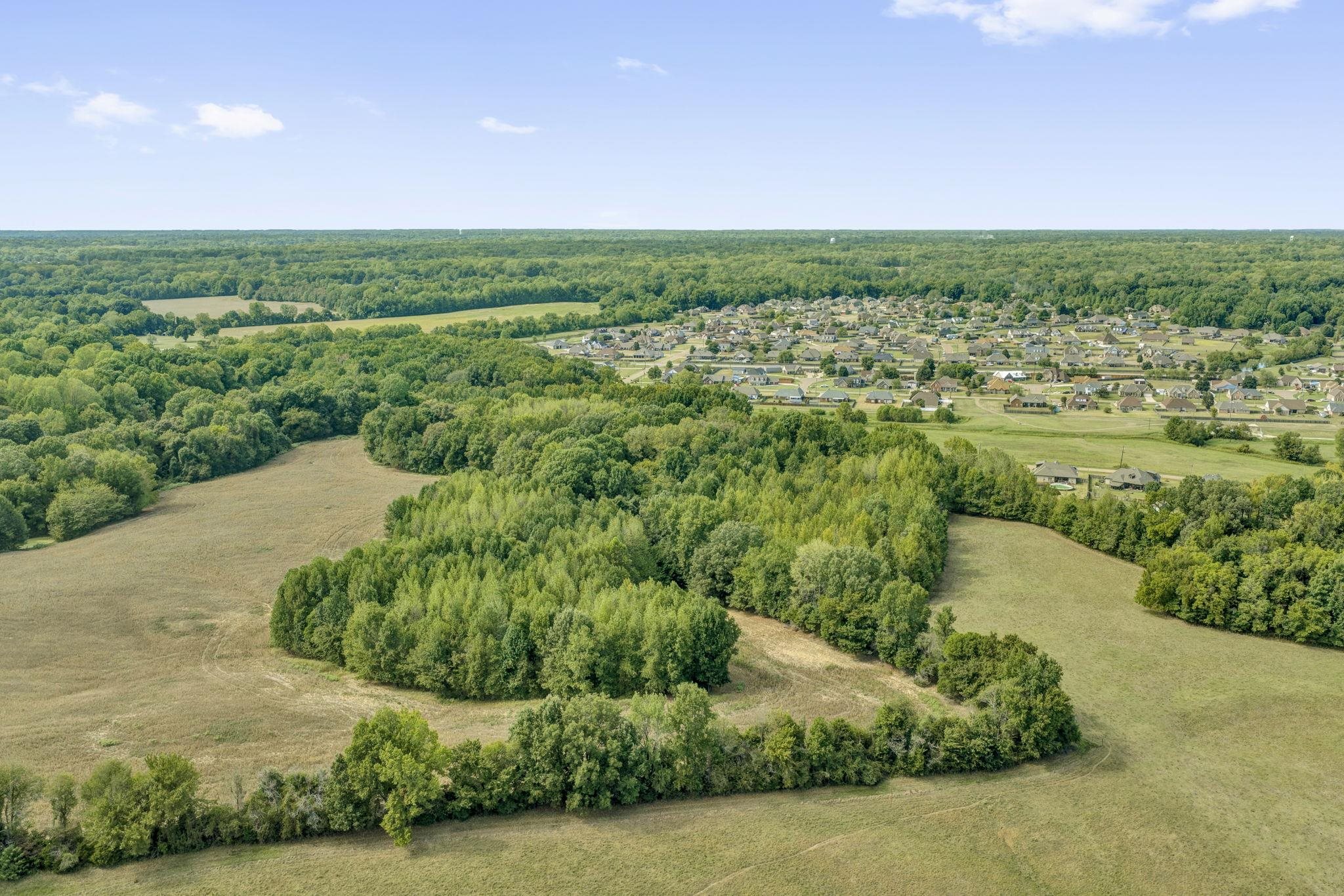 Aerial view of open fields and wooded land with a residential subdivision in the distance.