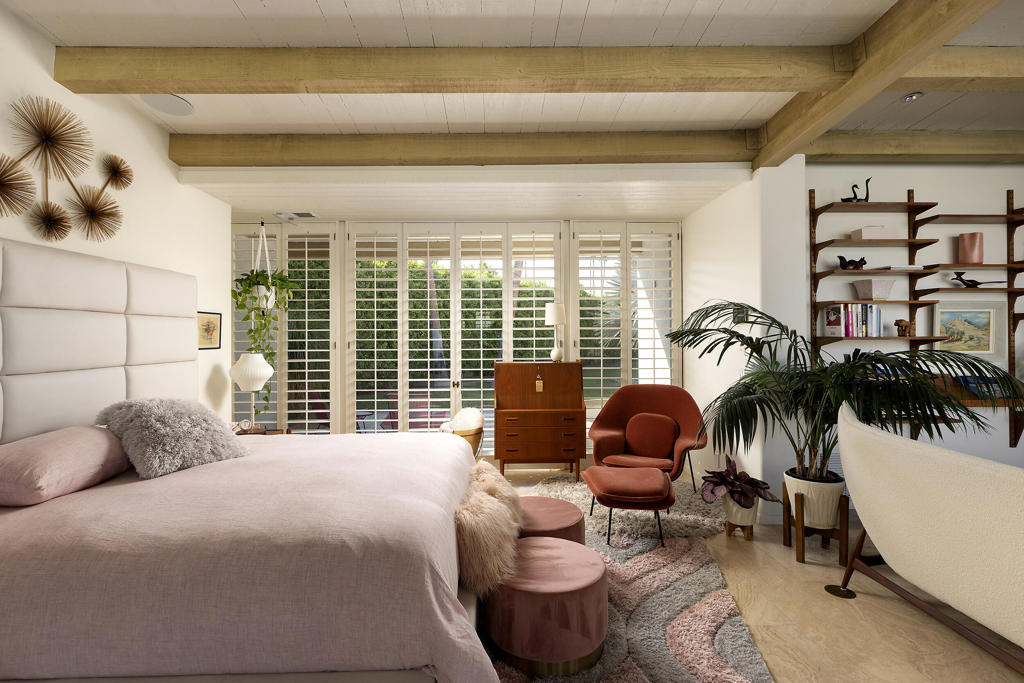 Bedroom with a large bed, pink seating, indoor plants, and white plantation shutters.