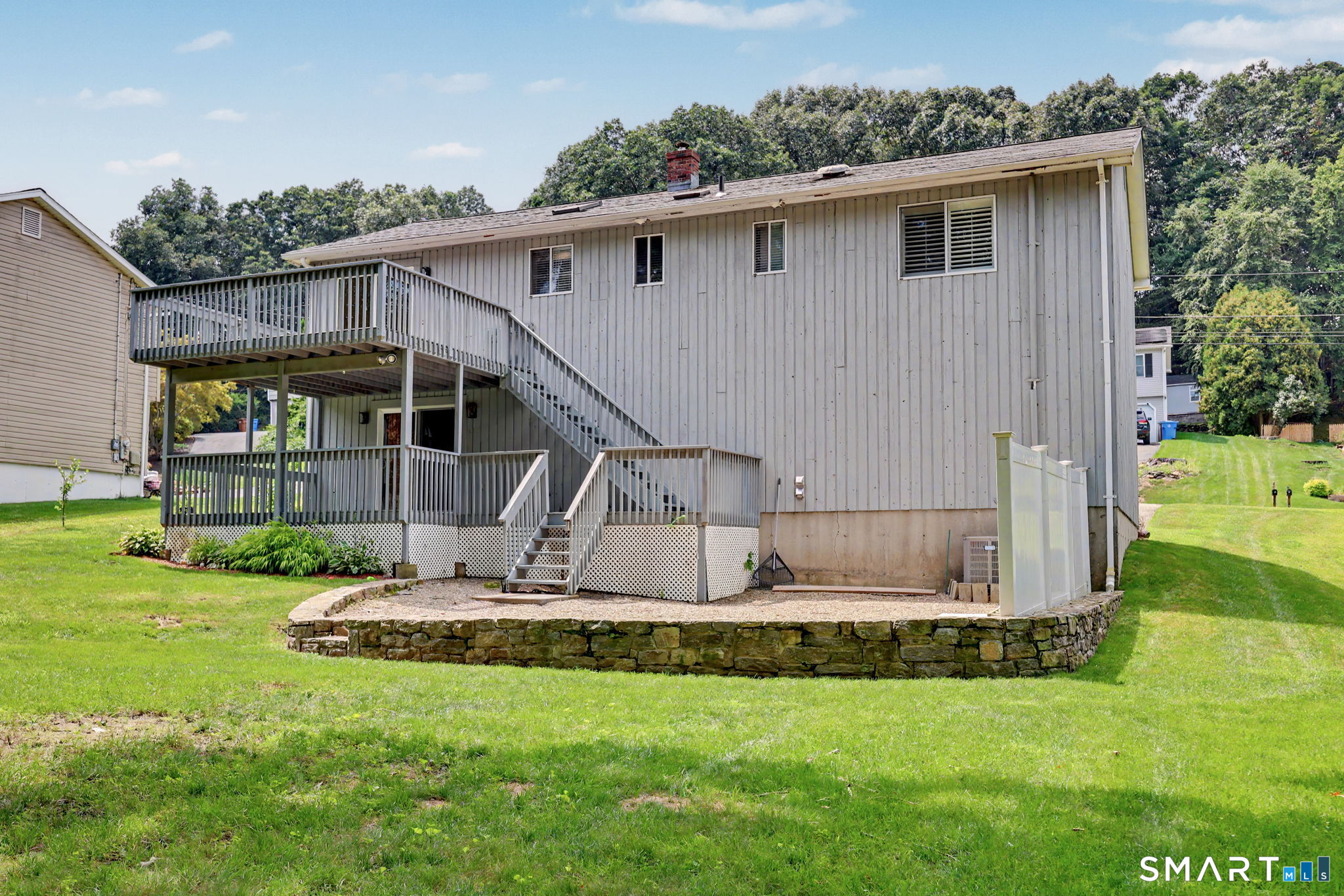 Rear of gray-sided home with a raised deck, exterior stairs, and stone-edged patio.