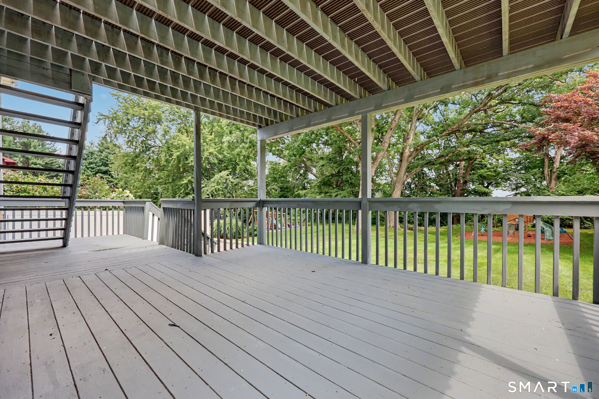 Covered gray deck overlooking a leafy backyard, with an exterior staircase and overhead beams.