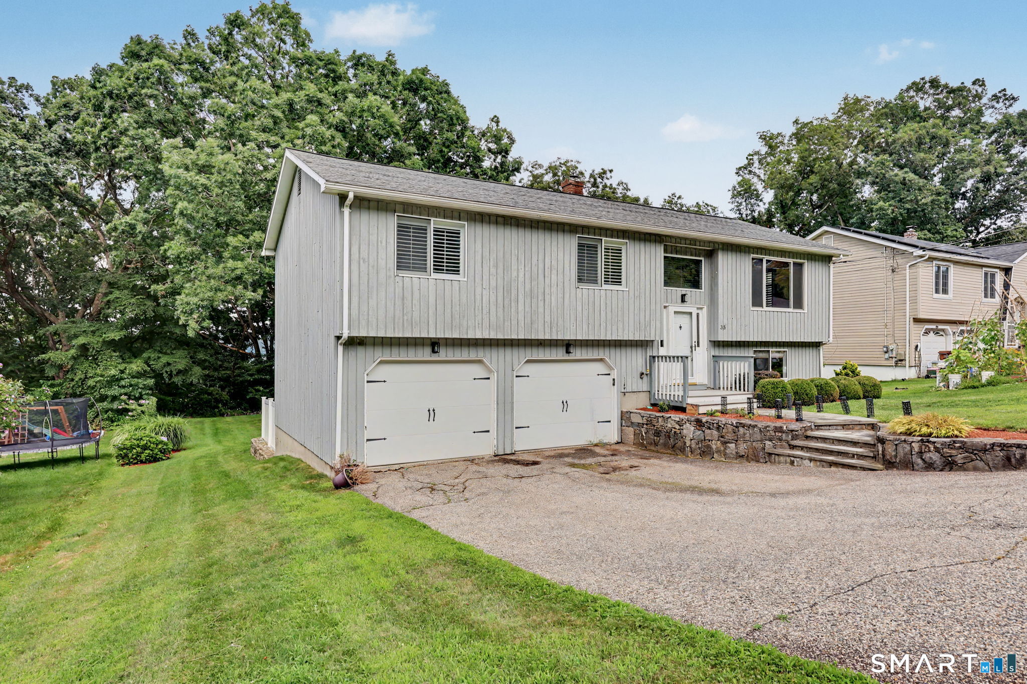Split-level home with two garage doors, a broad gravel driveway, lawn, and stone retaining wall.