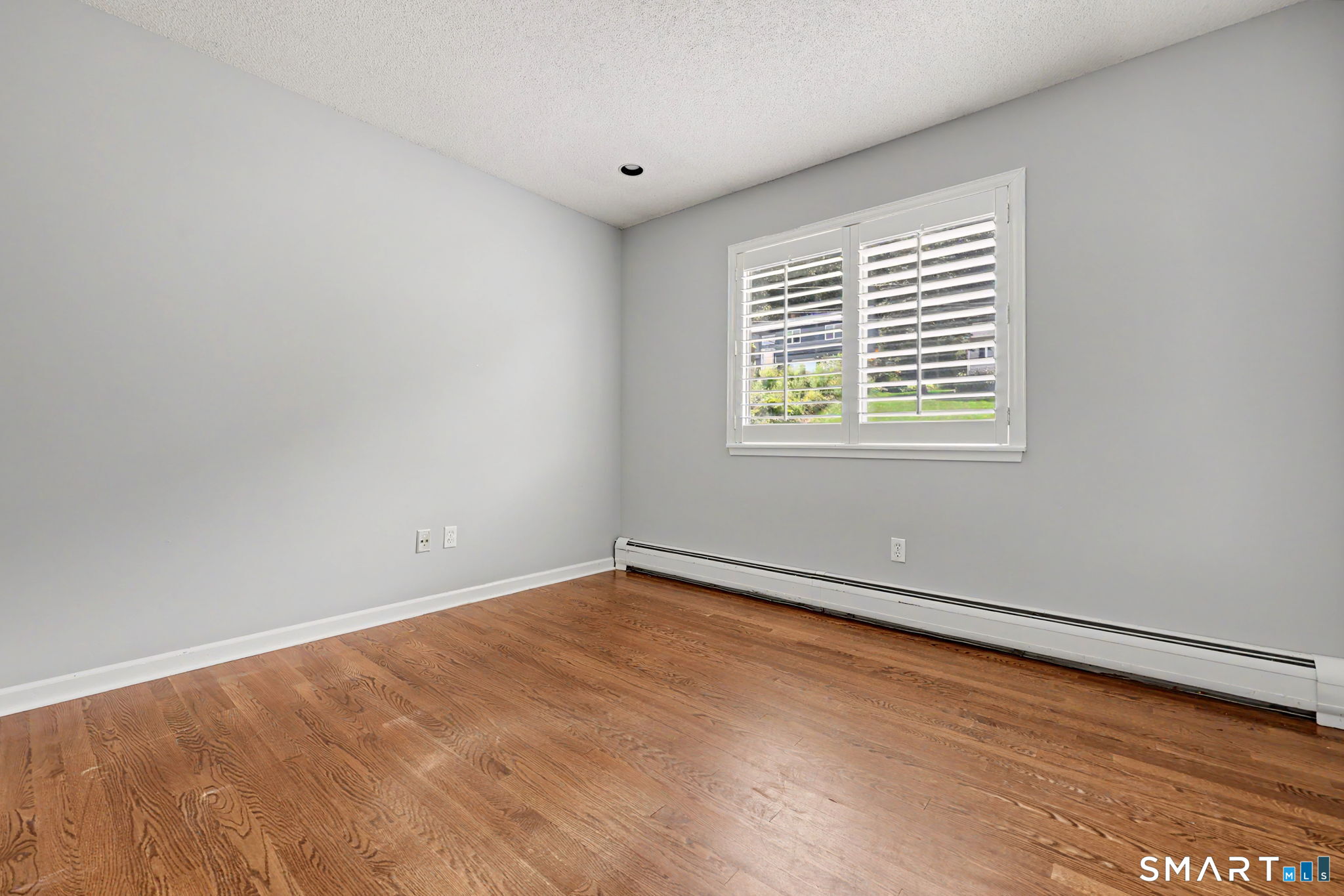 Empty room with hardwood floors, gray walls, shuttered window, and baseboard heater.
