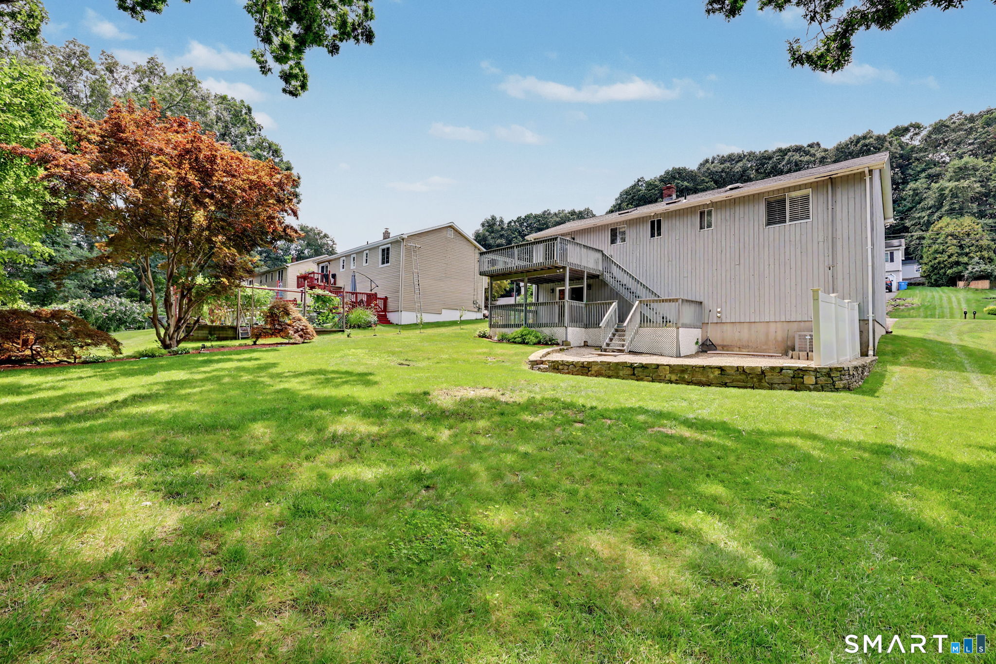 Rear of the home with a raised deck, exterior stairs, stone retaining wall, and broad lawn.