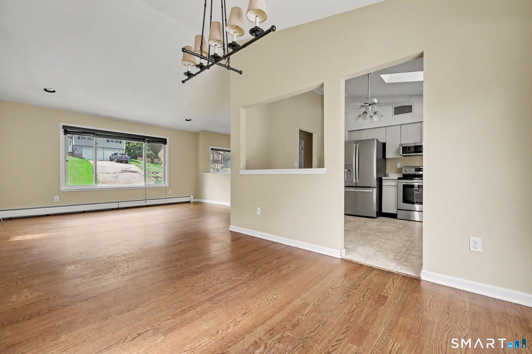 Empty living and dining area with hardwood floors, a large window, and an open kitchen with stainless steel appliances.