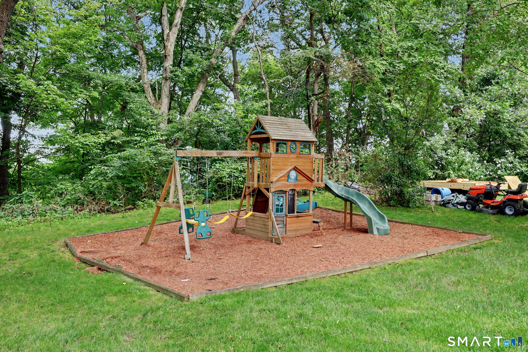 Backyard playset with swings and a green slide in a wooded lawn.