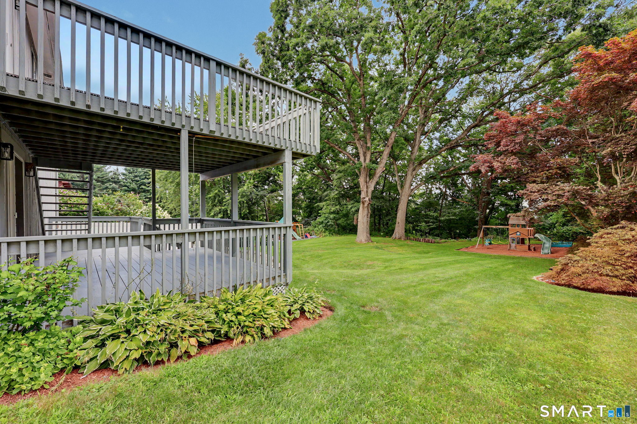 Backyard with two raised decks, a broad lawn, mature trees, and a children’s playset.