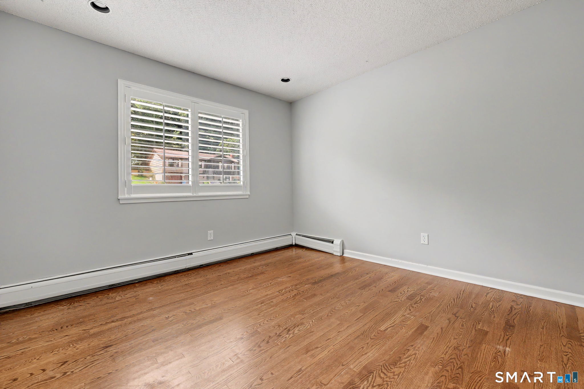 Empty room with hardwood floors, gray walls, and a window fitted with white plantation shutters.
