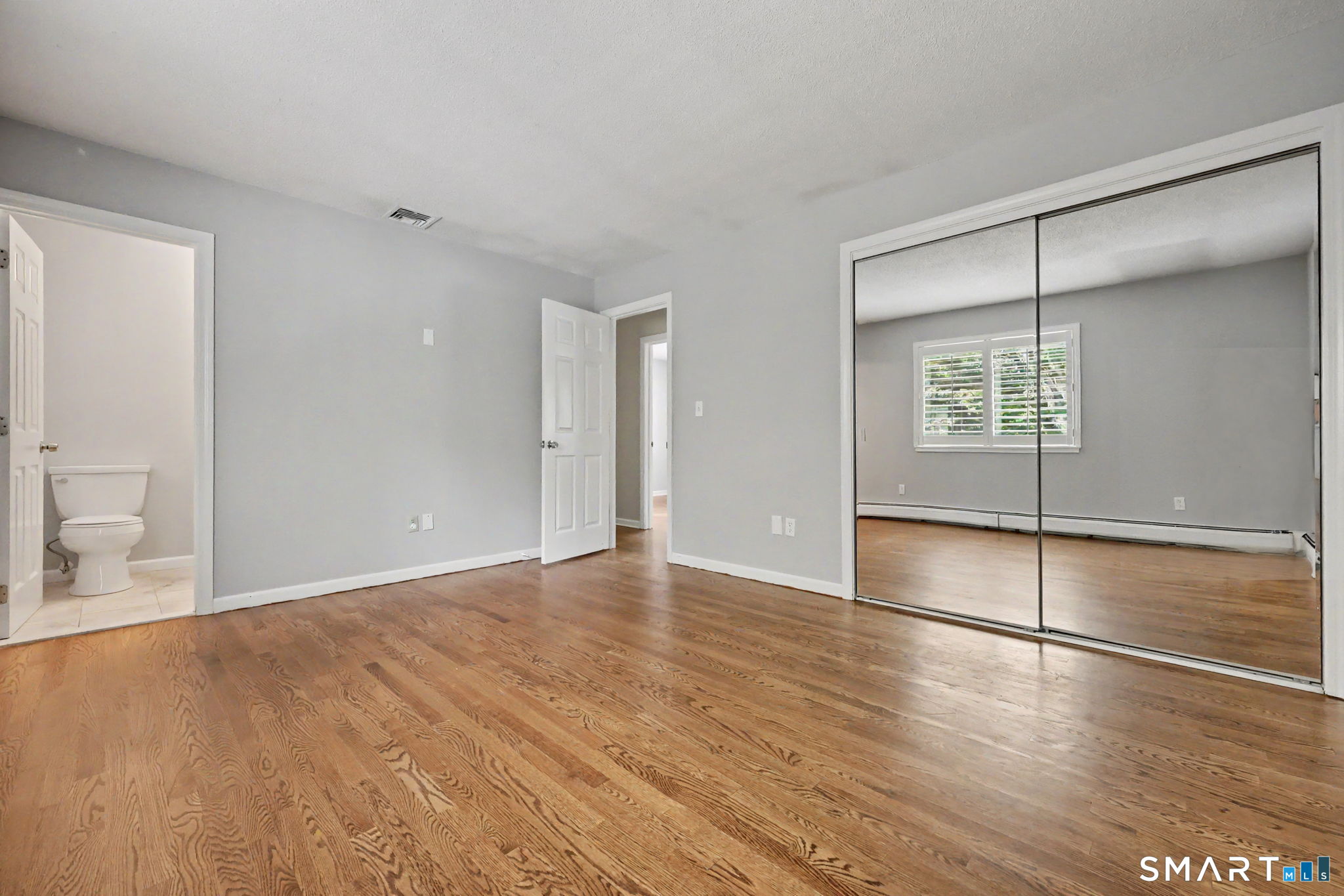 Empty bedroom with hardwood floors and mirrored closet doors, with an adjoining bathroom doorway.