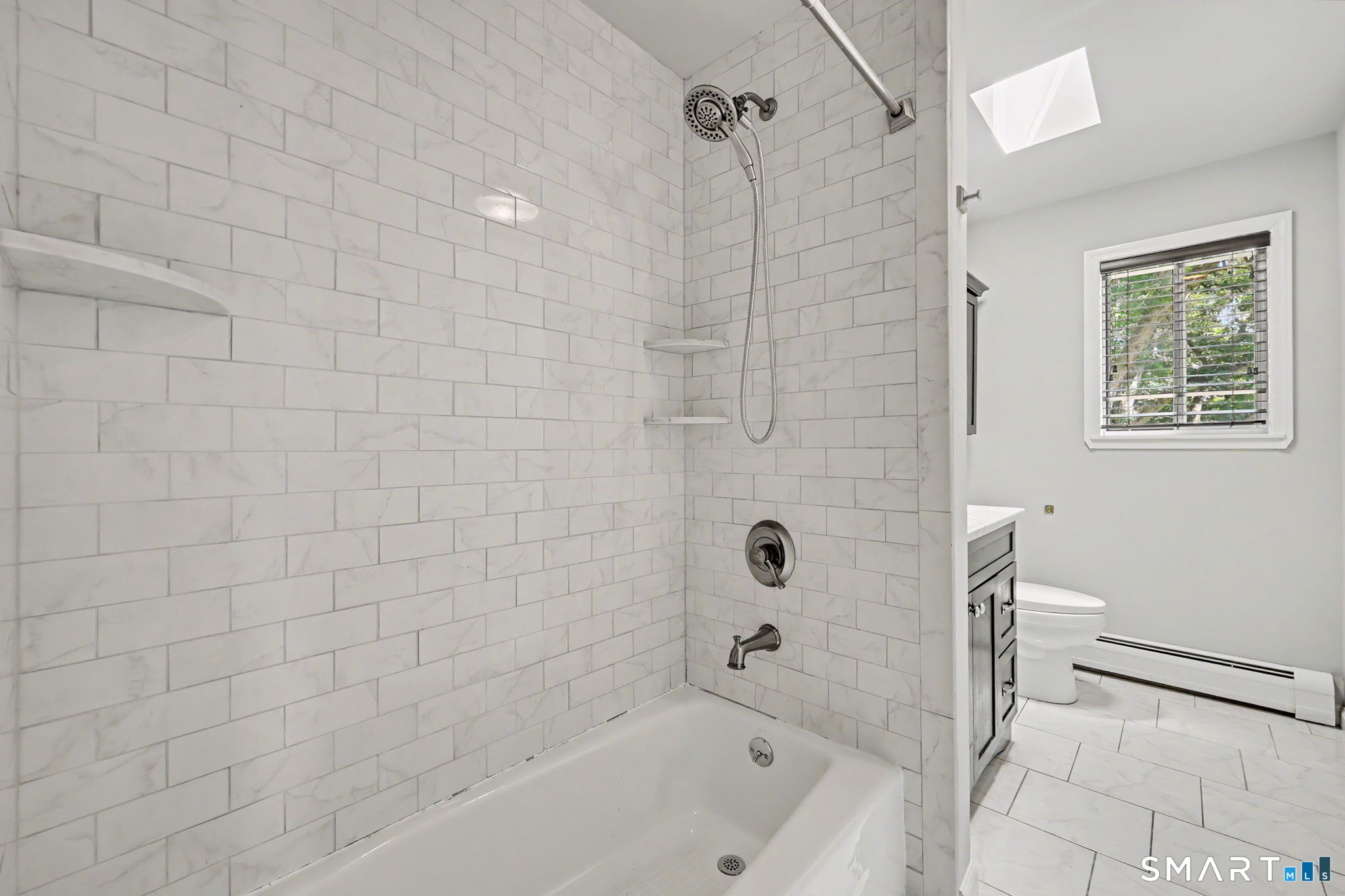 Bathroom with a marble-look tiled tub and shower beside a toilet and window.