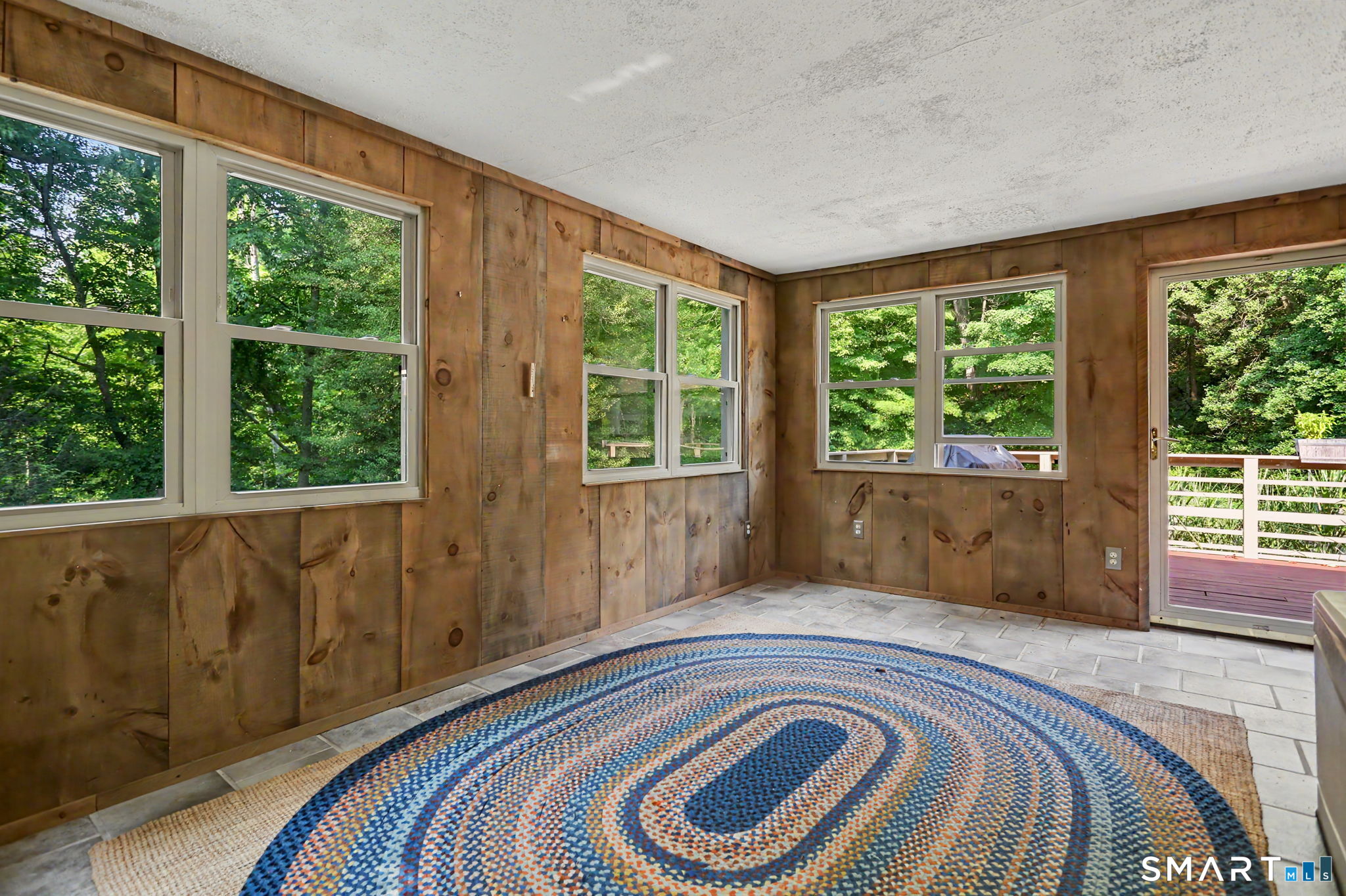 Sunroom with knotty wood walls, large leafy-view windows, tiled floor, and a round braided rug.