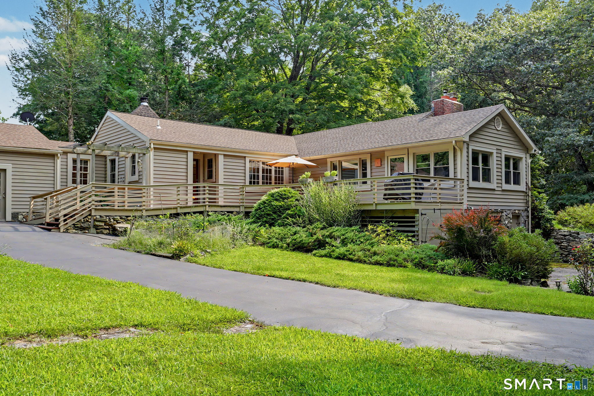 Single-story beige home with a broad raised deck and ramp, surrounded by lawn and mature trees.