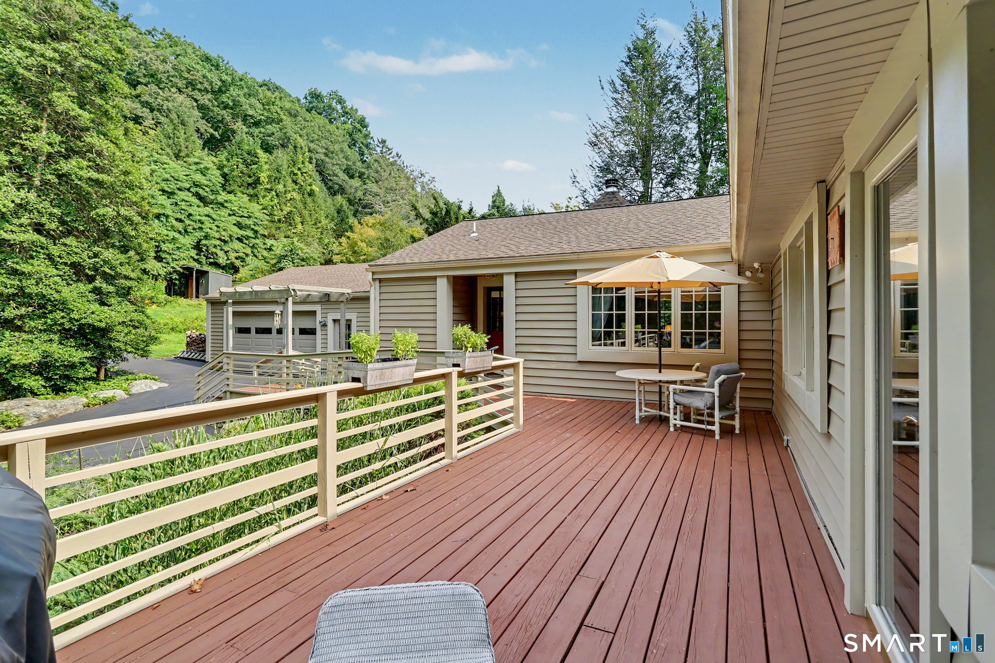 Spacious deck with umbrella-shaded seating, potted plants, and a wooded backdrop.