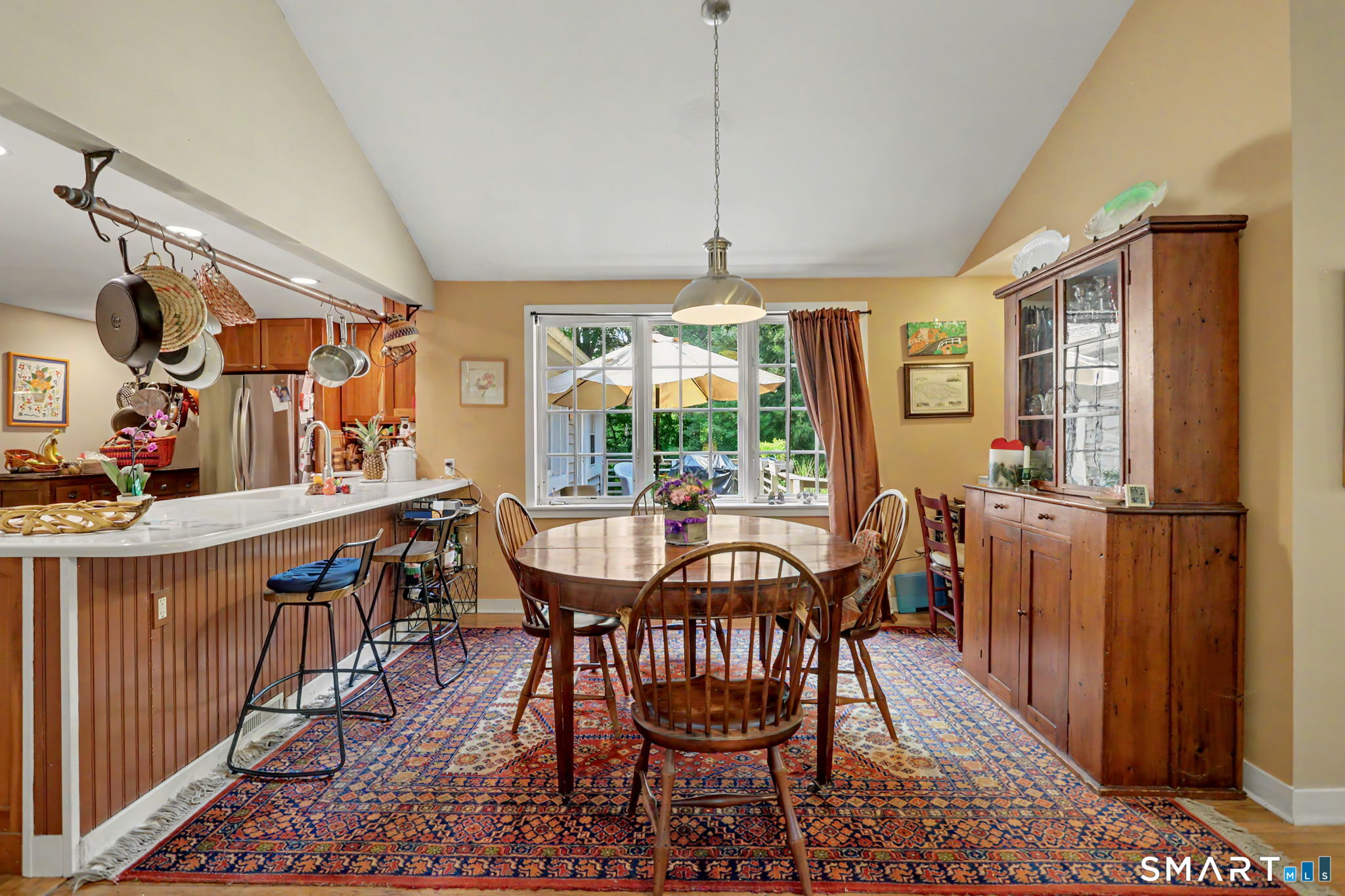 Kitchen and dining area with a white island, hanging cookware, and a round wooden table by large windows.