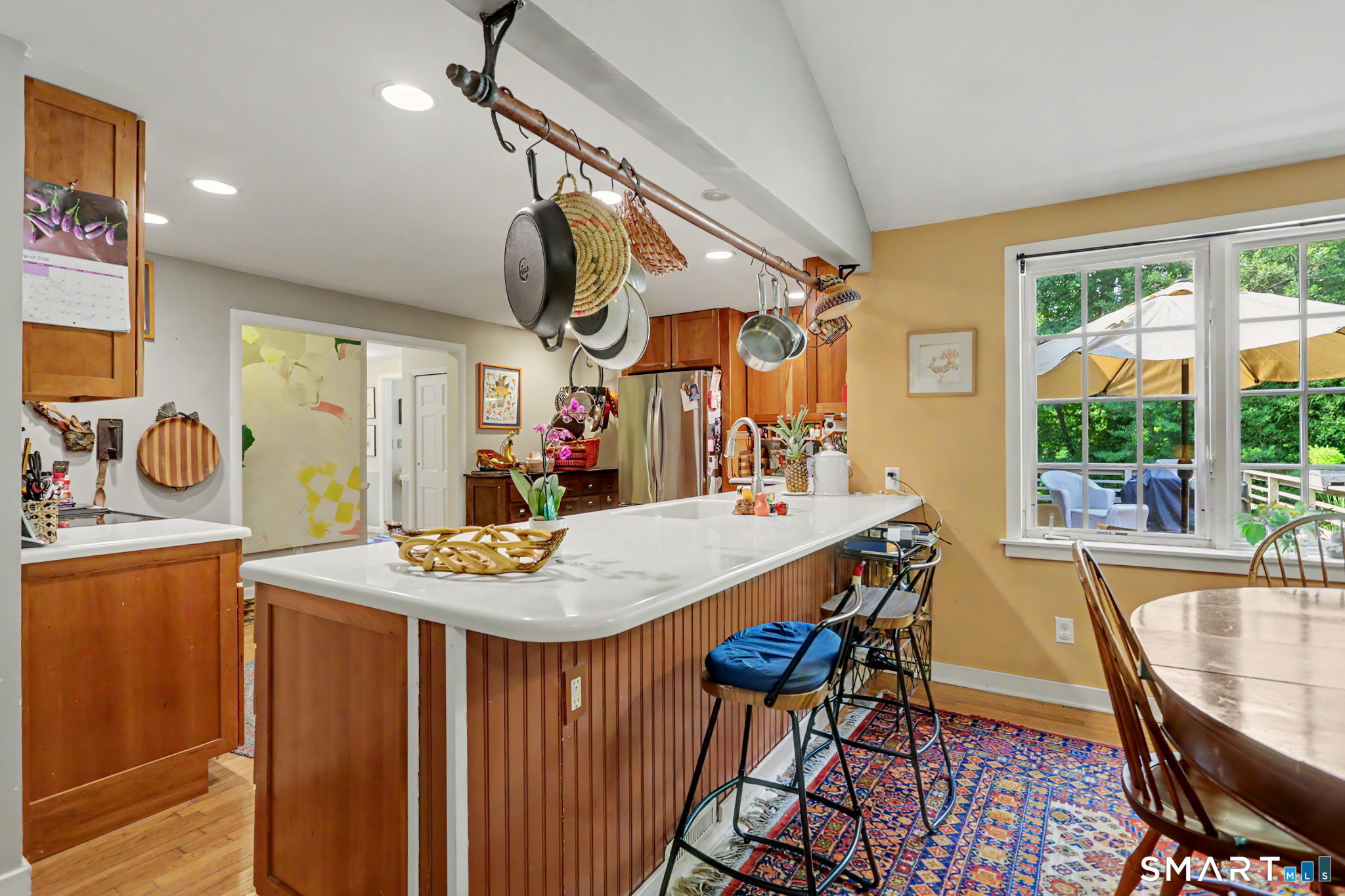 Kitchen with a large white island, hanging cookware, warm wood cabinetry, and a window overlooking a leafy yard.