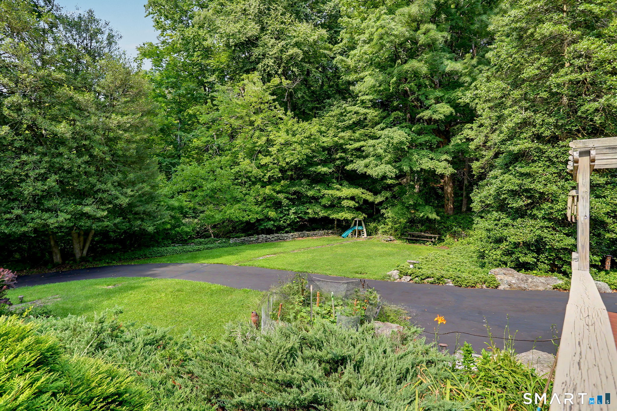 Landscaped backyard with a broad lawn, mature trees, paved drive, and a small playset.