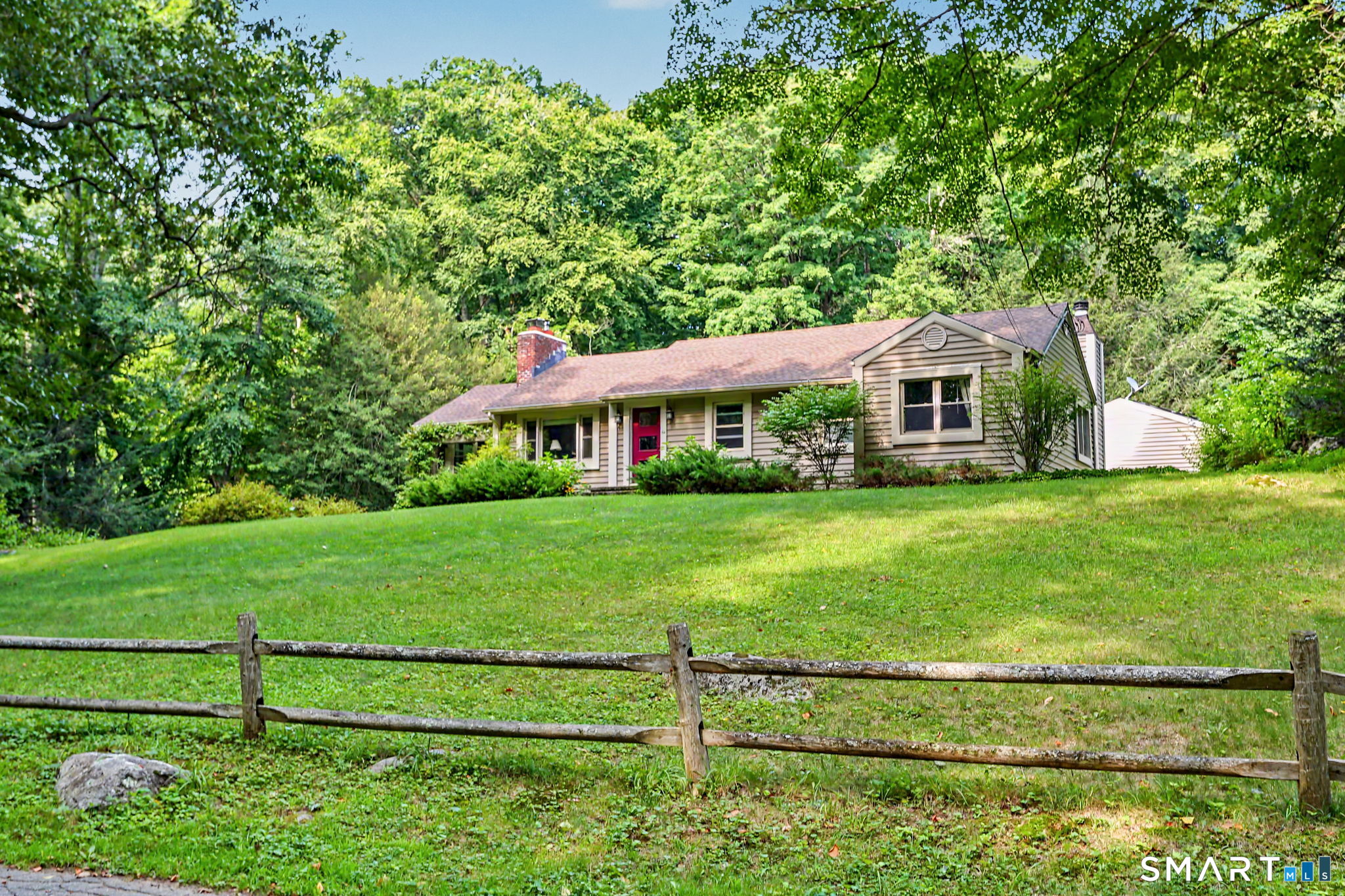 Single-story house with a red front door, broad lawn, split-rail fence, and wooded surroundings.