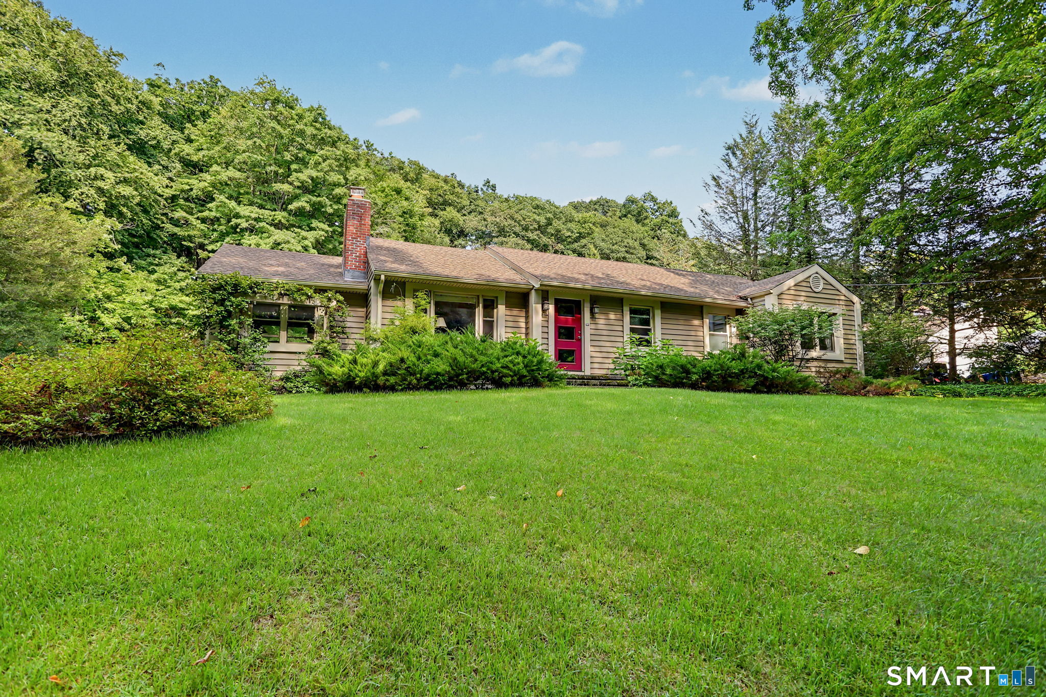 Single-story home with a red front door, brick chimney, and broad lawn surrounded by trees.