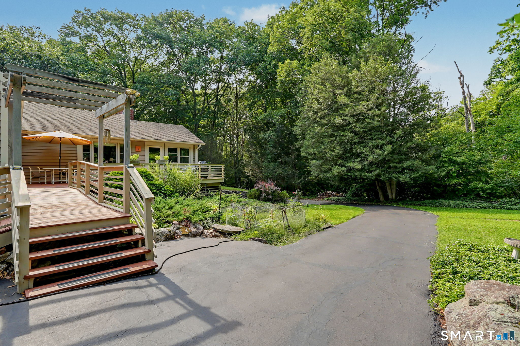 House with a raised wooden deck and pergola beside a landscaped driveway and mature trees.